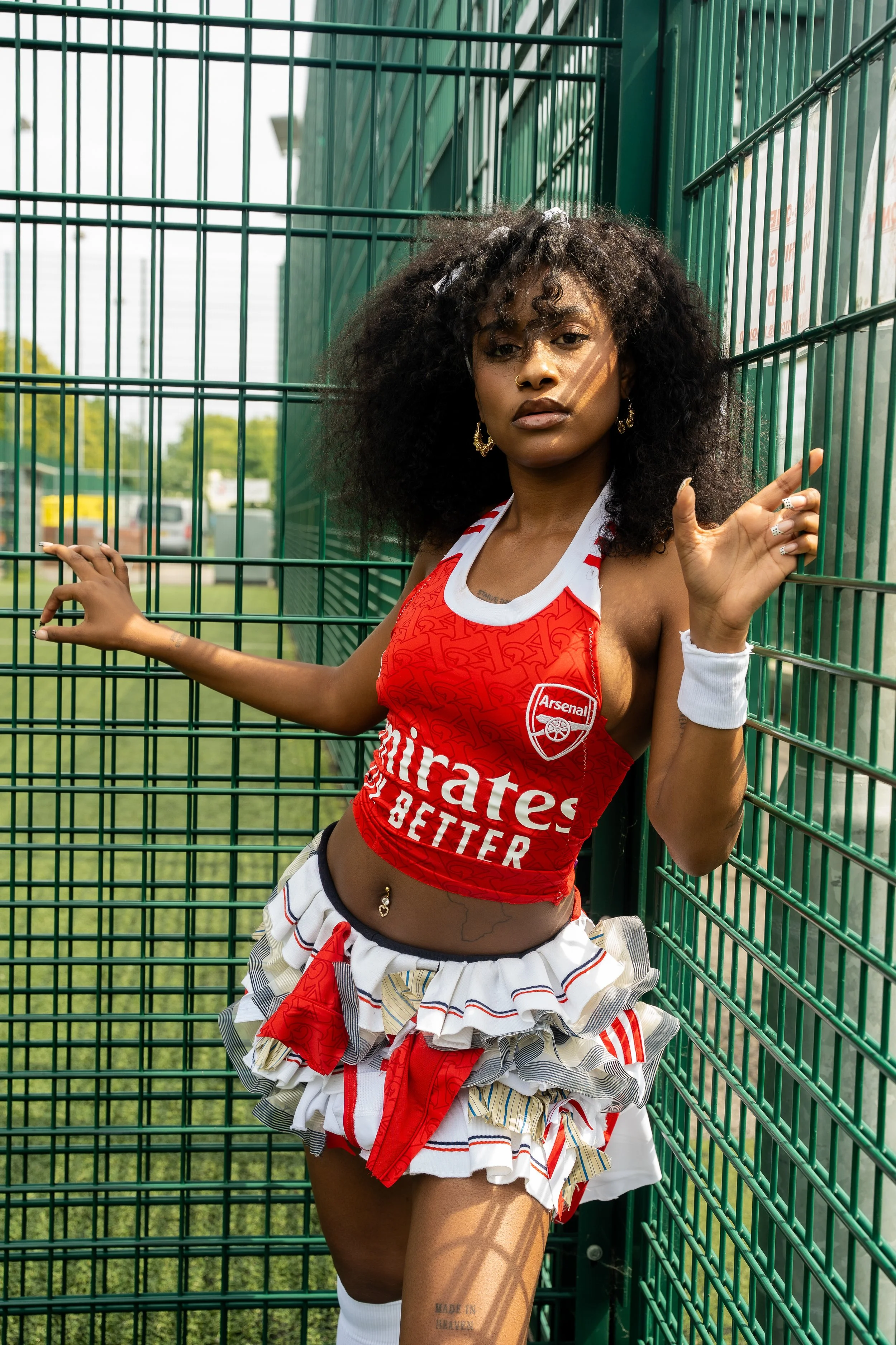 Young woman in Arsenal football club cheerleader outfit with red top, ruffled skirt, and white accessories, standing inside a green metal fence at an outdoor sports field.