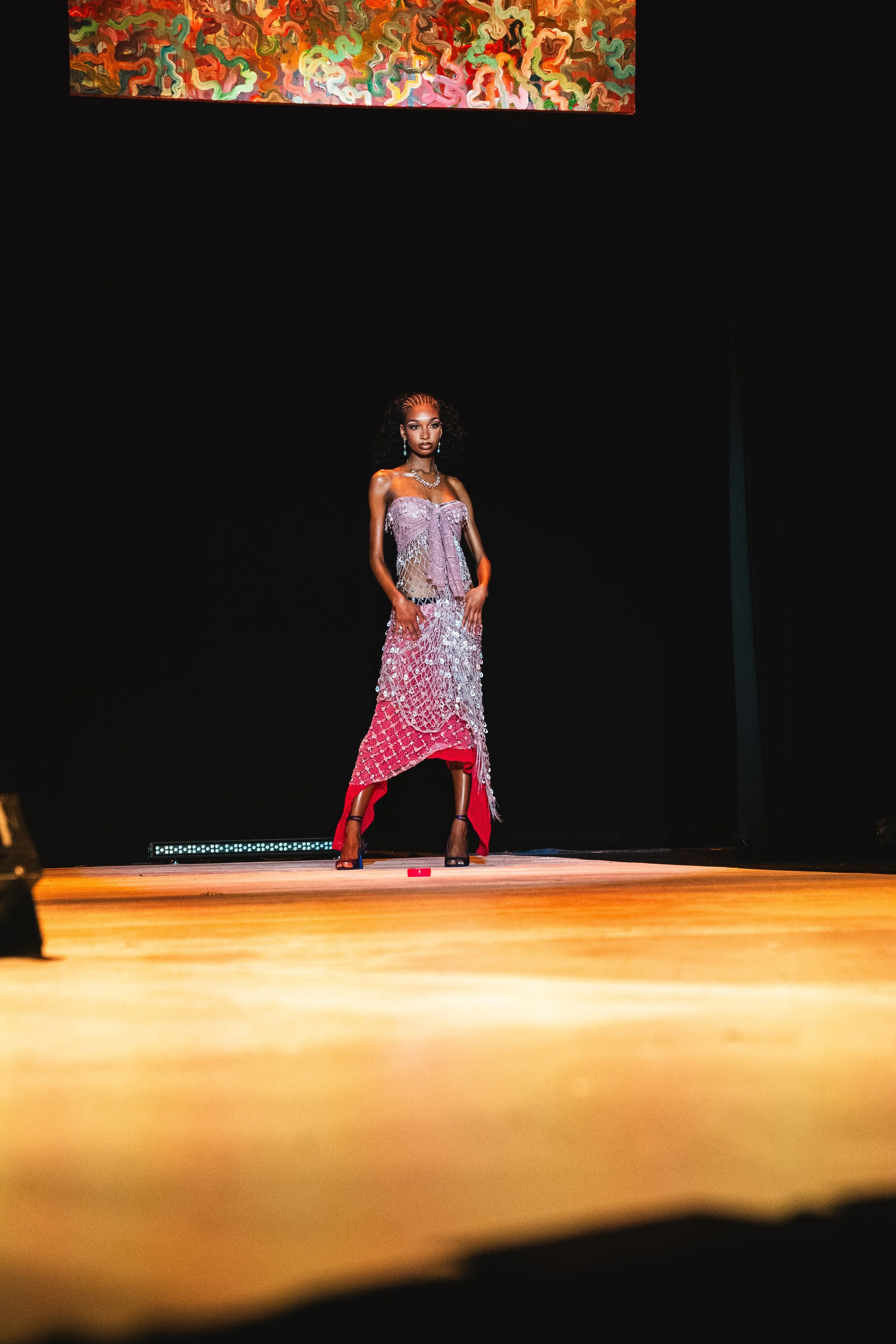 A woman stands on a stage wearing a colorful dress with intricate patterns, with a dark background and a piece of abstract art hanging above her.