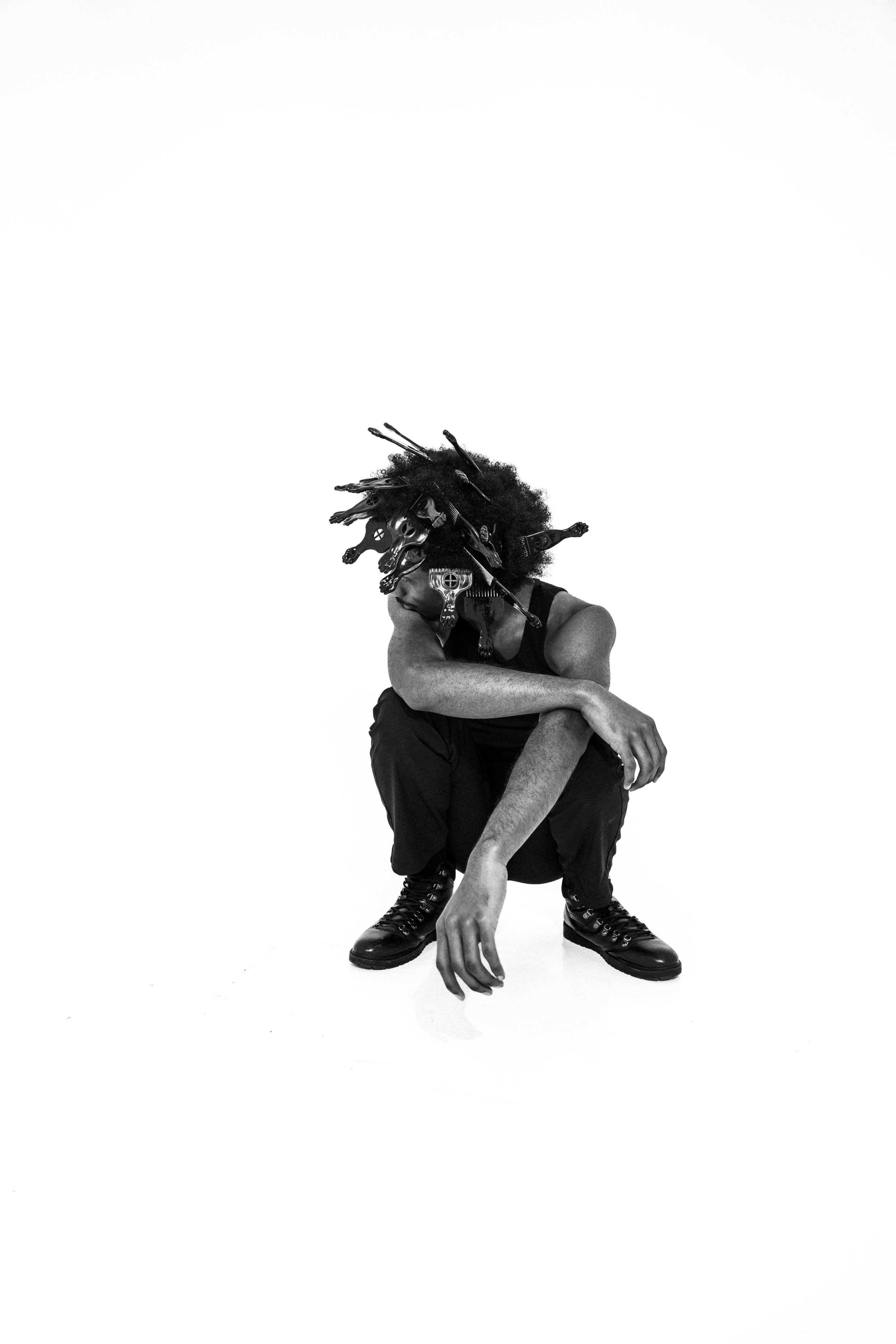 A person squatting with their head covered in an artful arrangement of animal skulls and antlers, wearing dark clothing and boots, against a plain white background.