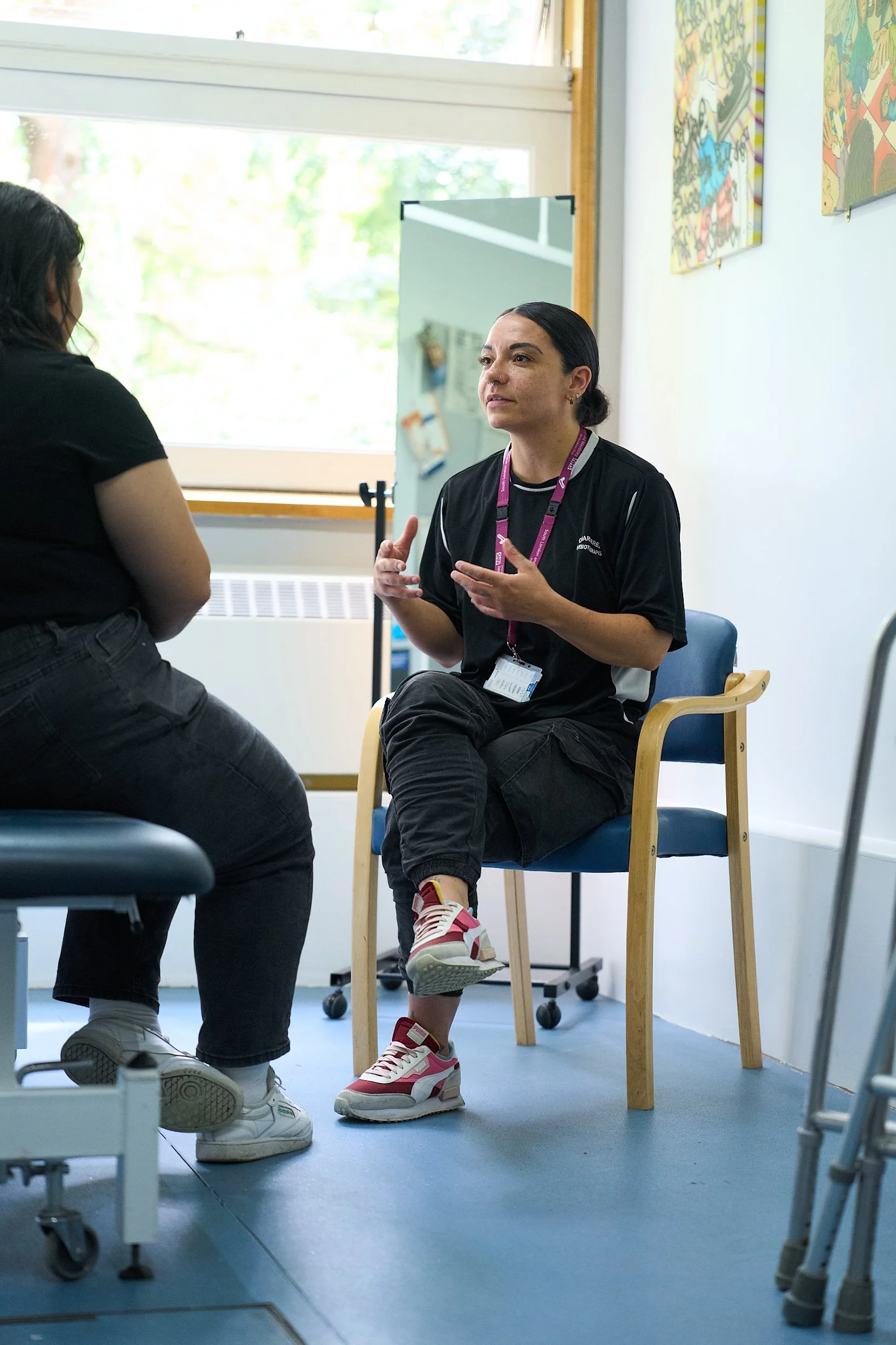 Two women engaged in conversation inside a brightly lit room with large window and colorful artwork on the wall.