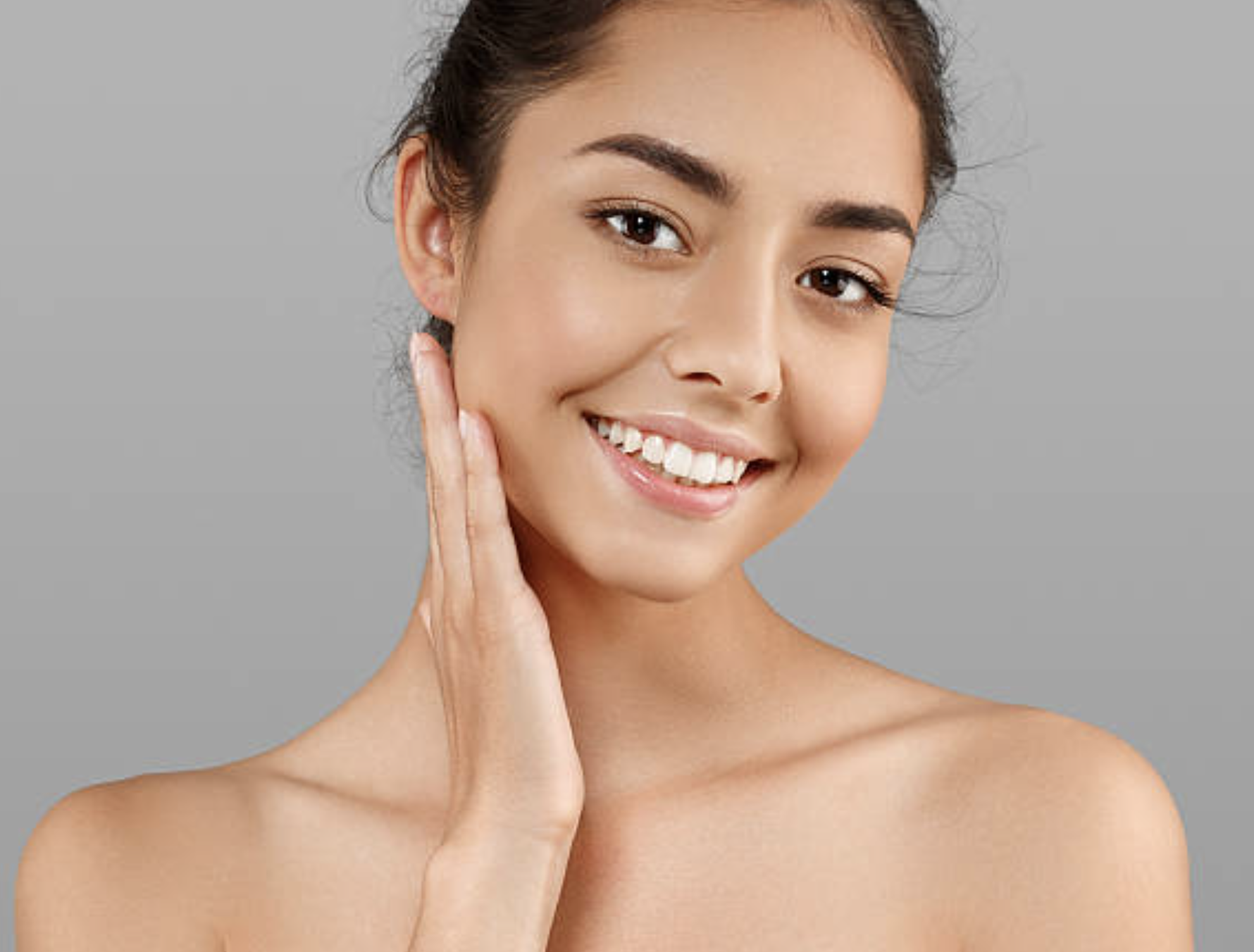 A young woman with medium skin tone, dark hair tied back, smiling, touching her face with her left hand, bare shoulders, against a plain gray background.