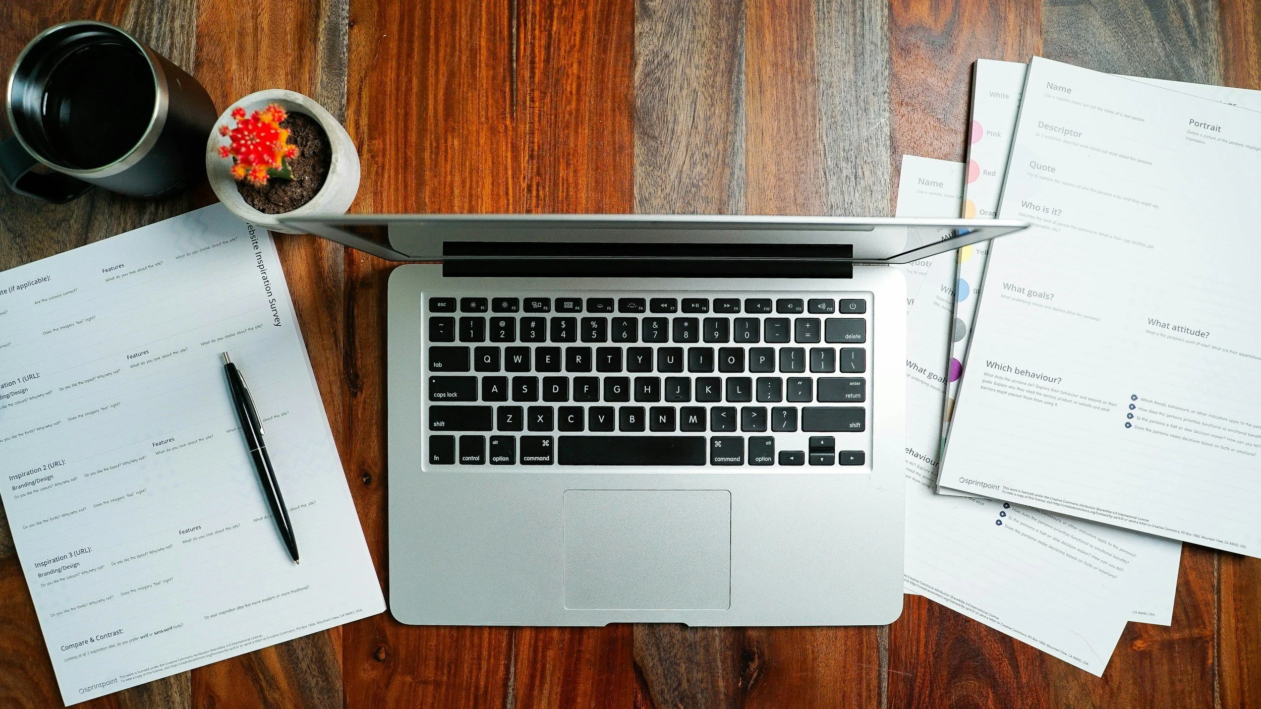 A laptop on a wooden desk with scattered sheets of paper, a pen, a black coffee mug, and a small potted cactus.