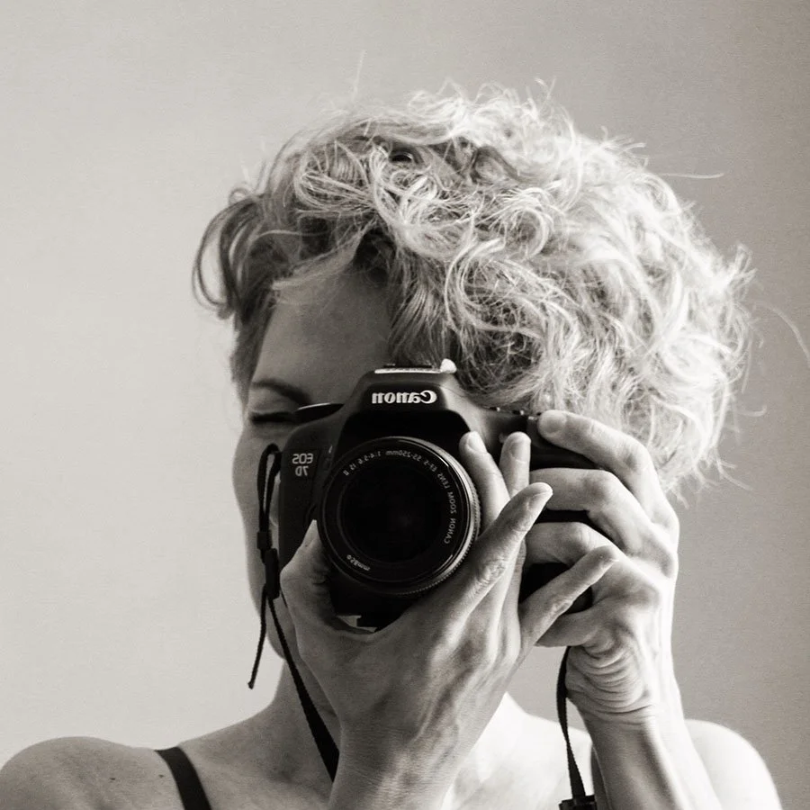 A woman with curly, short blonde hair taking a photo with a Canon camera, mostly covering her face, in black and white.