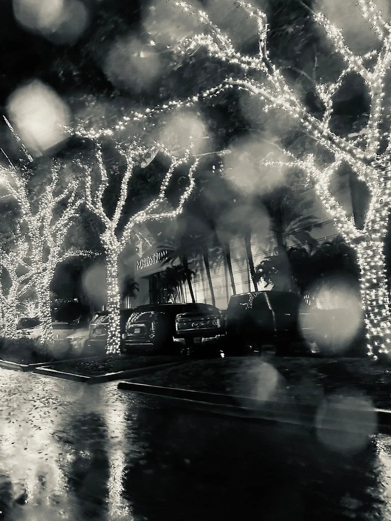 Nighttime street scene of a shopping center with trees wrapped in string lights, parked cars, and raindrops on the camera lens creating a blurred and wet effect.