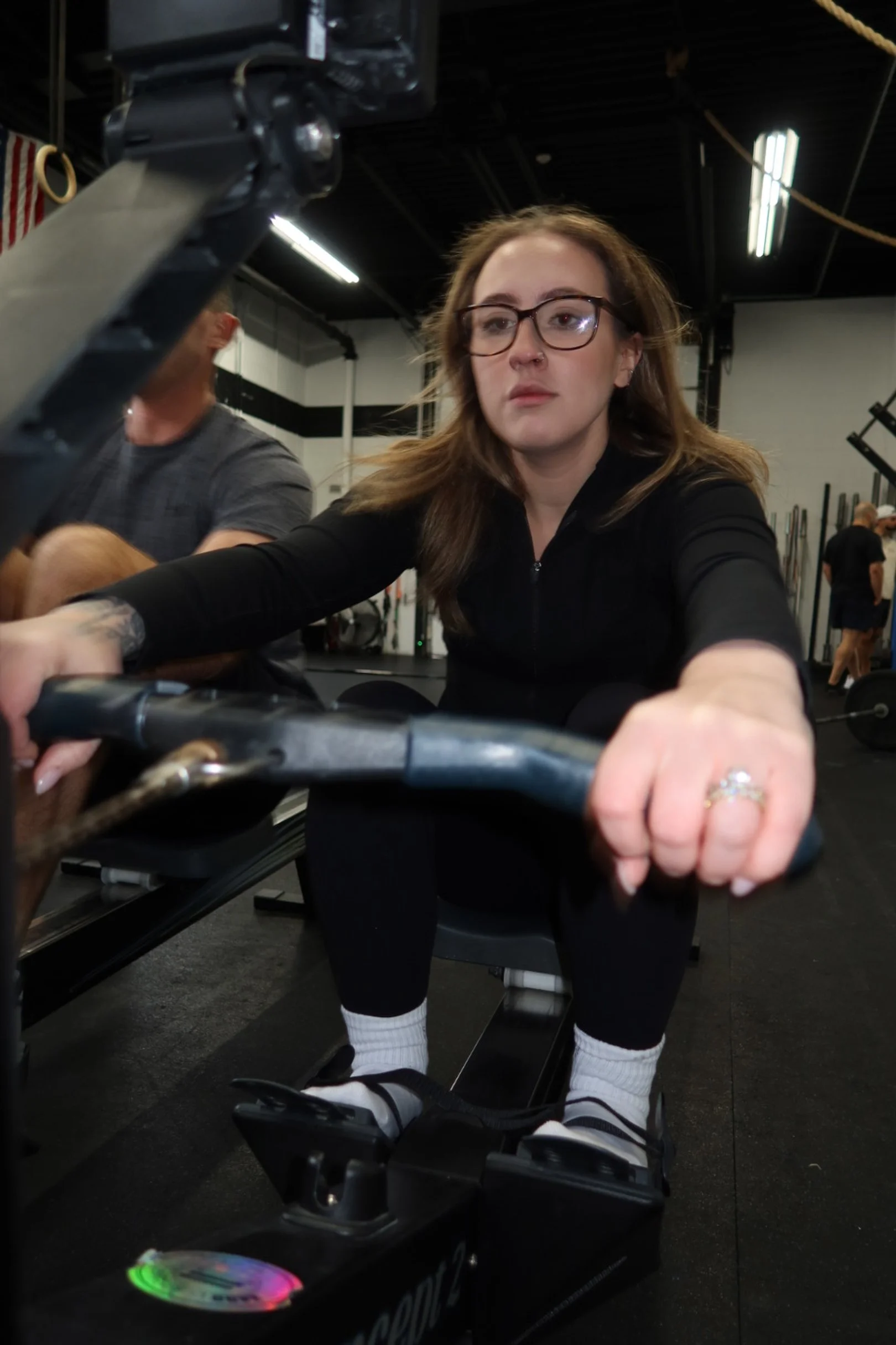 A woman with glasses and long brown hair using a rowing machine in a gym, with a focused expression on her face.
