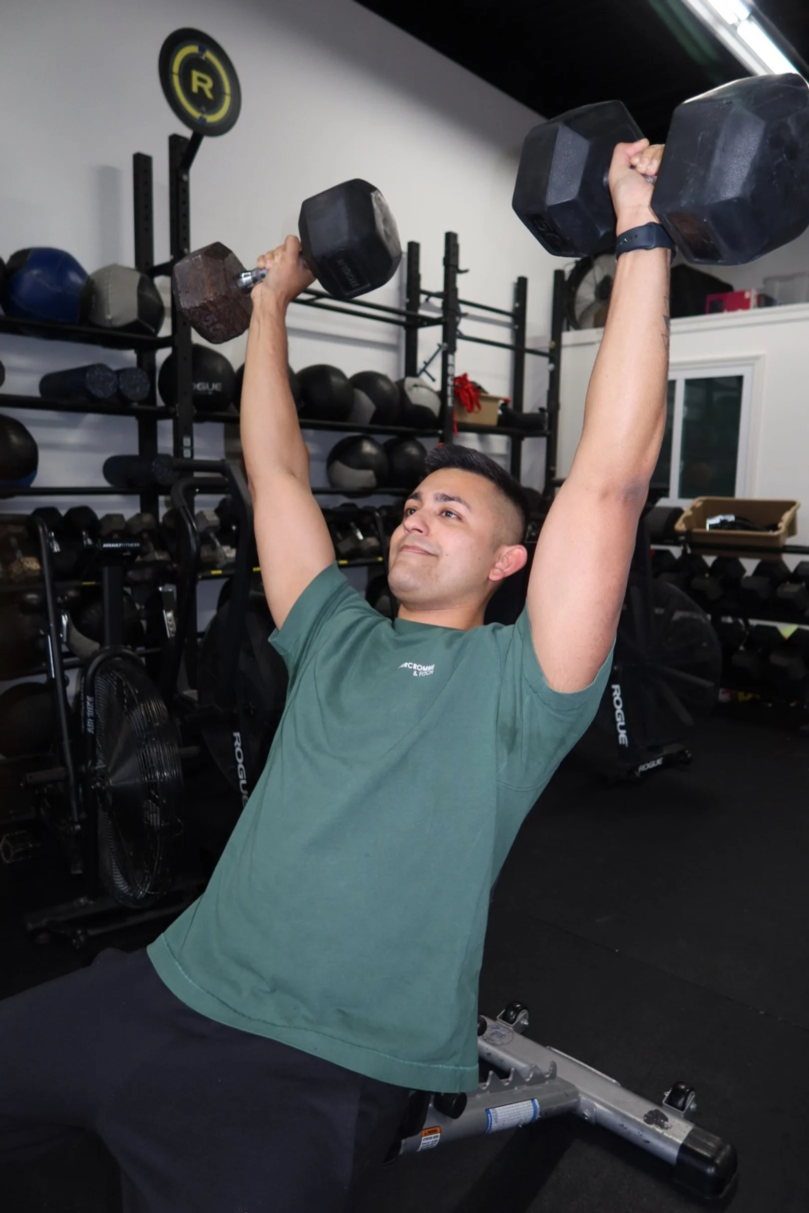 Man doing an overhead dumbbell press in a gym, seated on a workout bench, with gym equipment and kettlebells on shelves behind him.