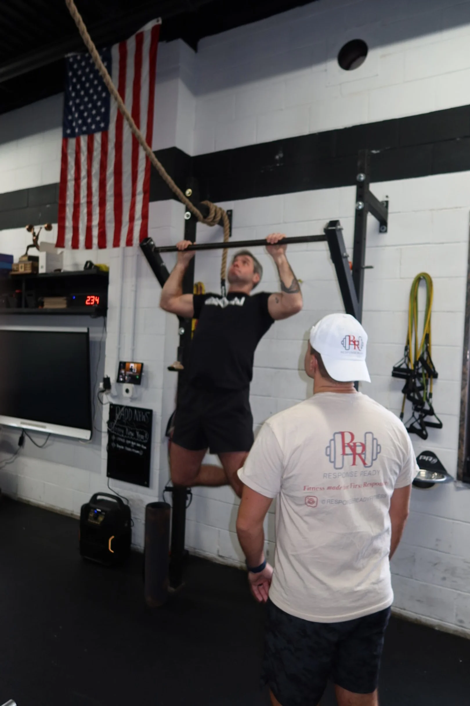 A man doing pull-ups on a pull-up bar in a gym, with another man watching. An American flag and workout equipment are visible in the background.