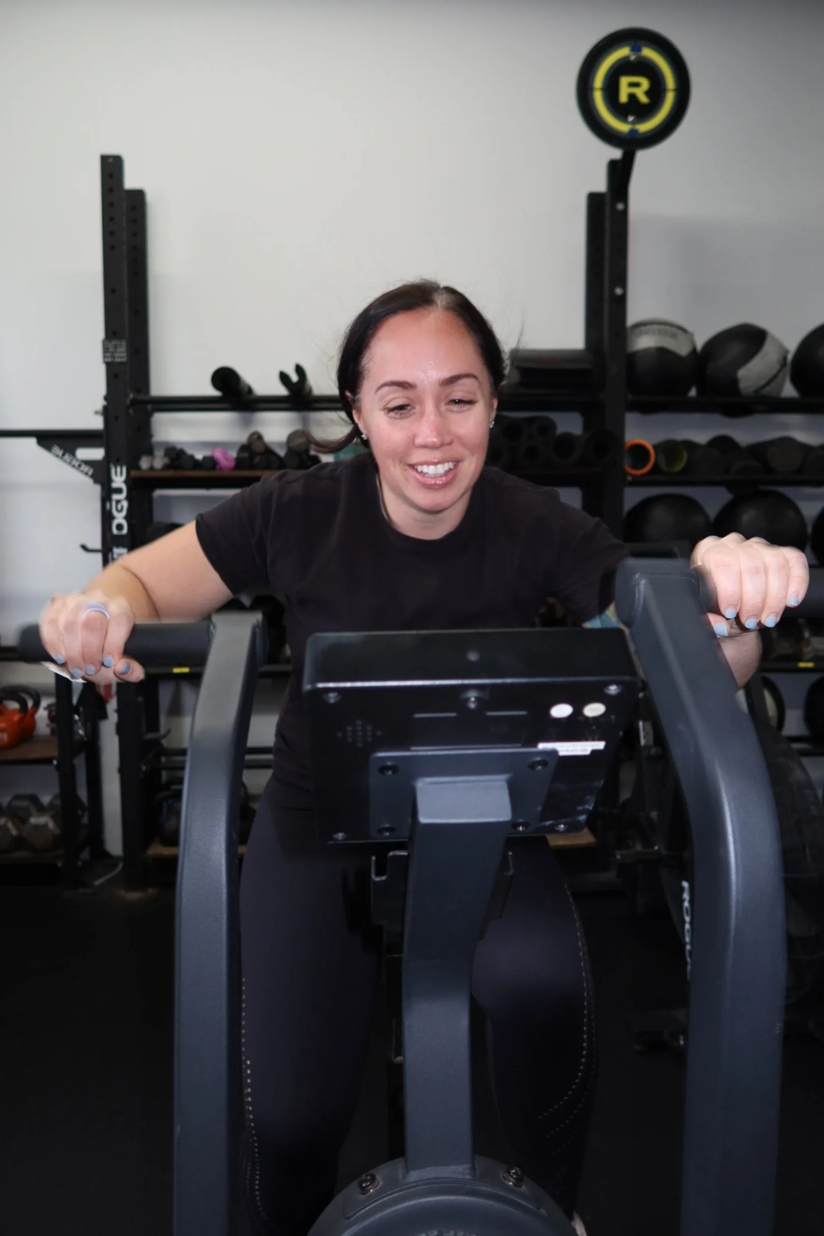 Woman smiling while using an exercise bike in a gym with workout equipment in the background.