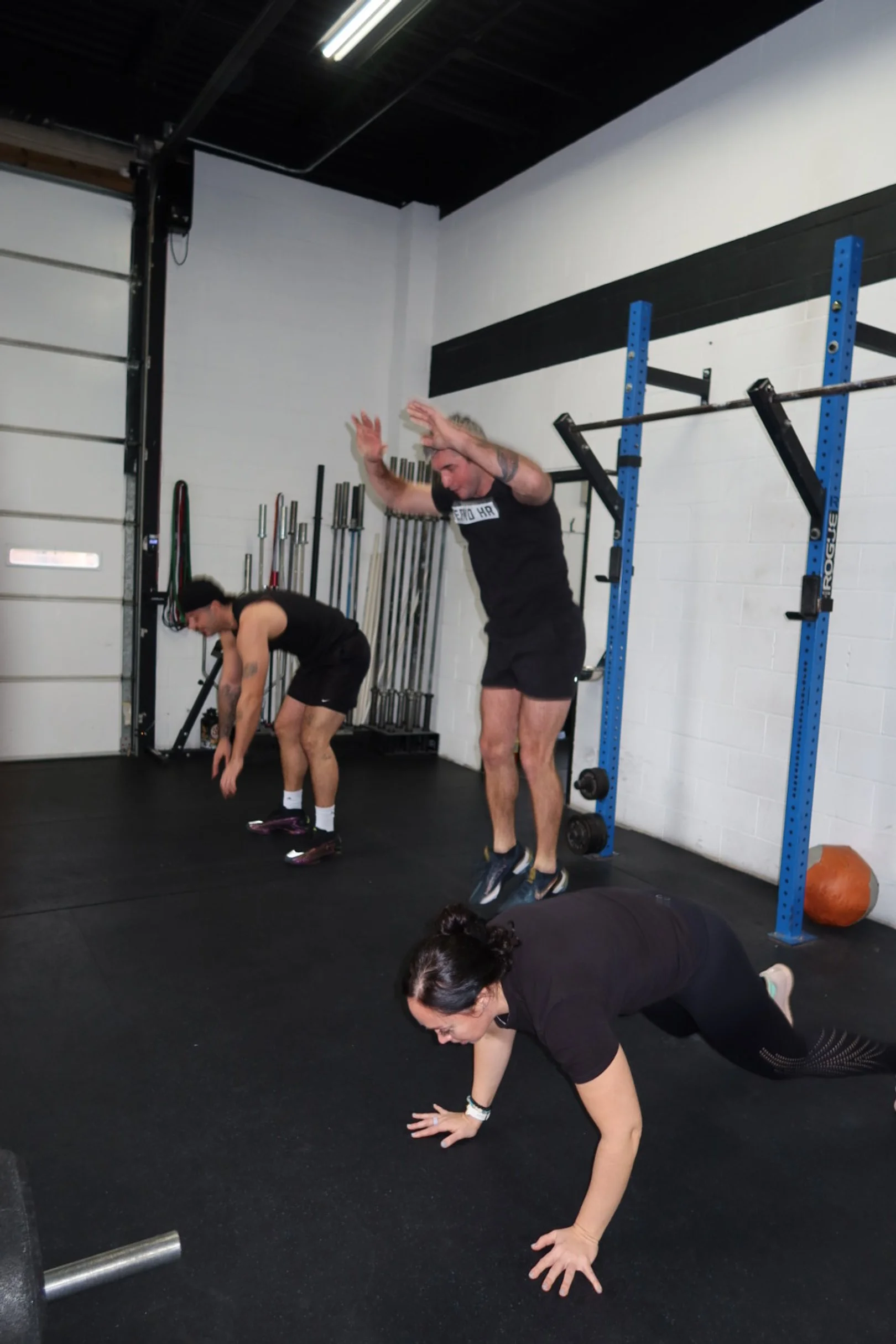 Three people perform exercises on black flooring in a gym, with a woman in front doing a push-up, a man in the middle jumping, and a woman in the back bending forward.