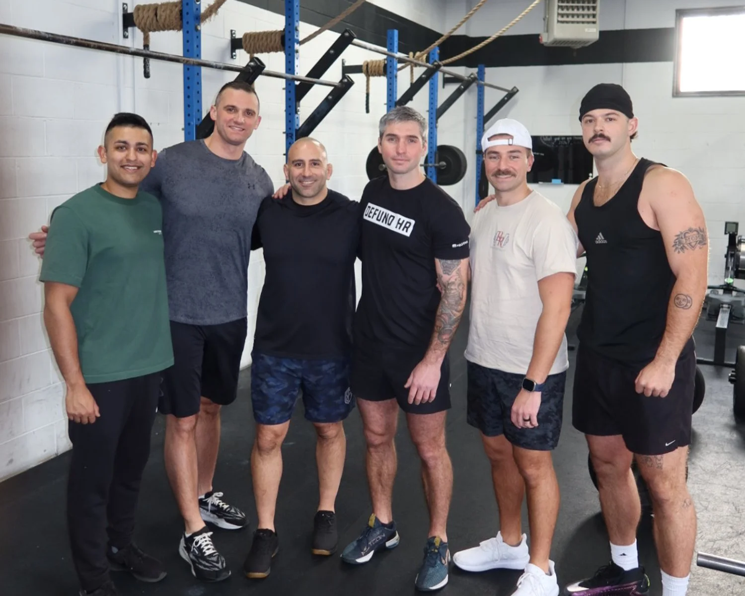 Group of six men standing together in a gym, smiling, with fitness equipment and a white brick wall in the background.