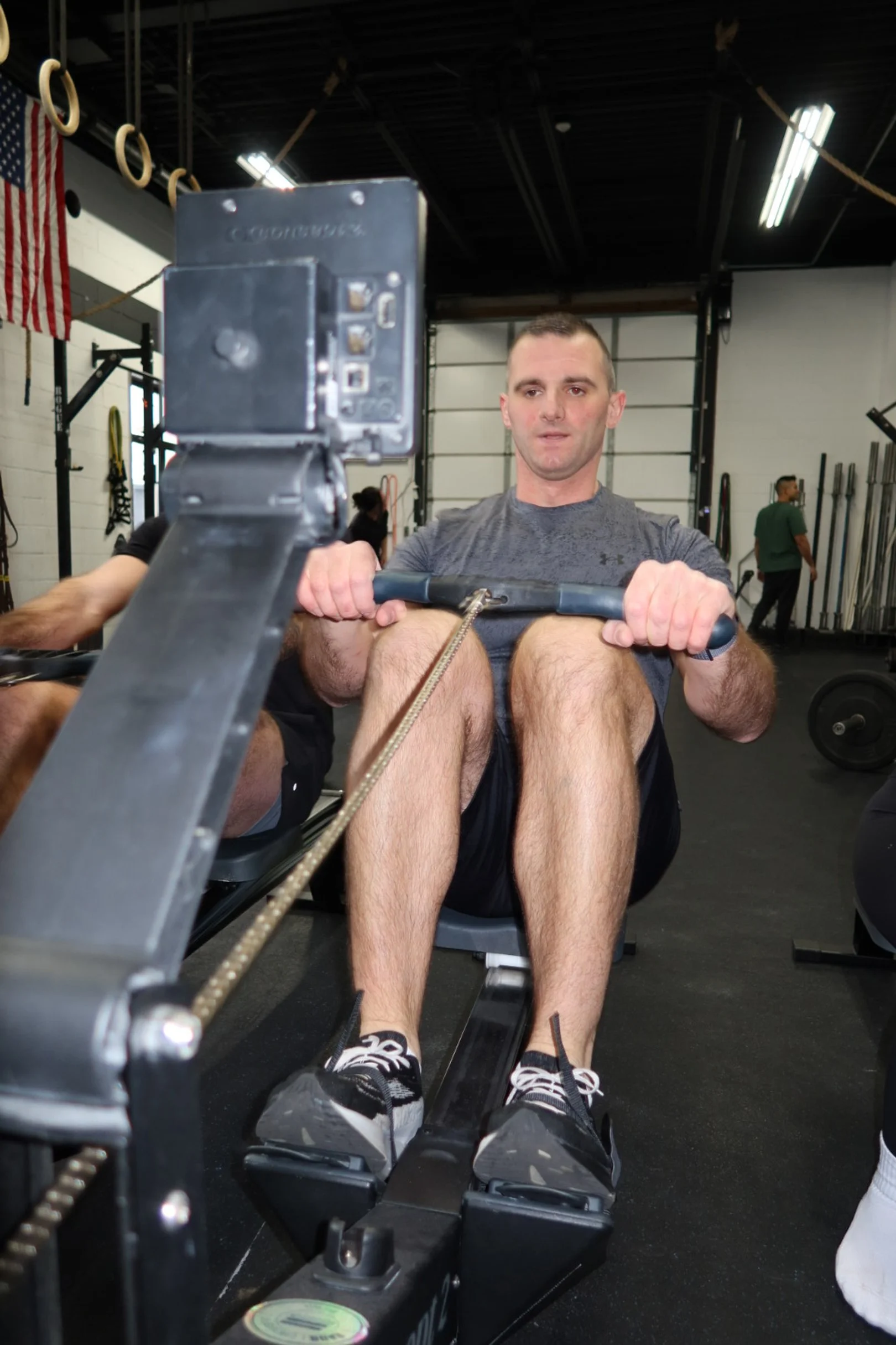 Man using a rowing machine in a gym with black flooring, American flag hanging on the wall, and other gym equipment in the background.