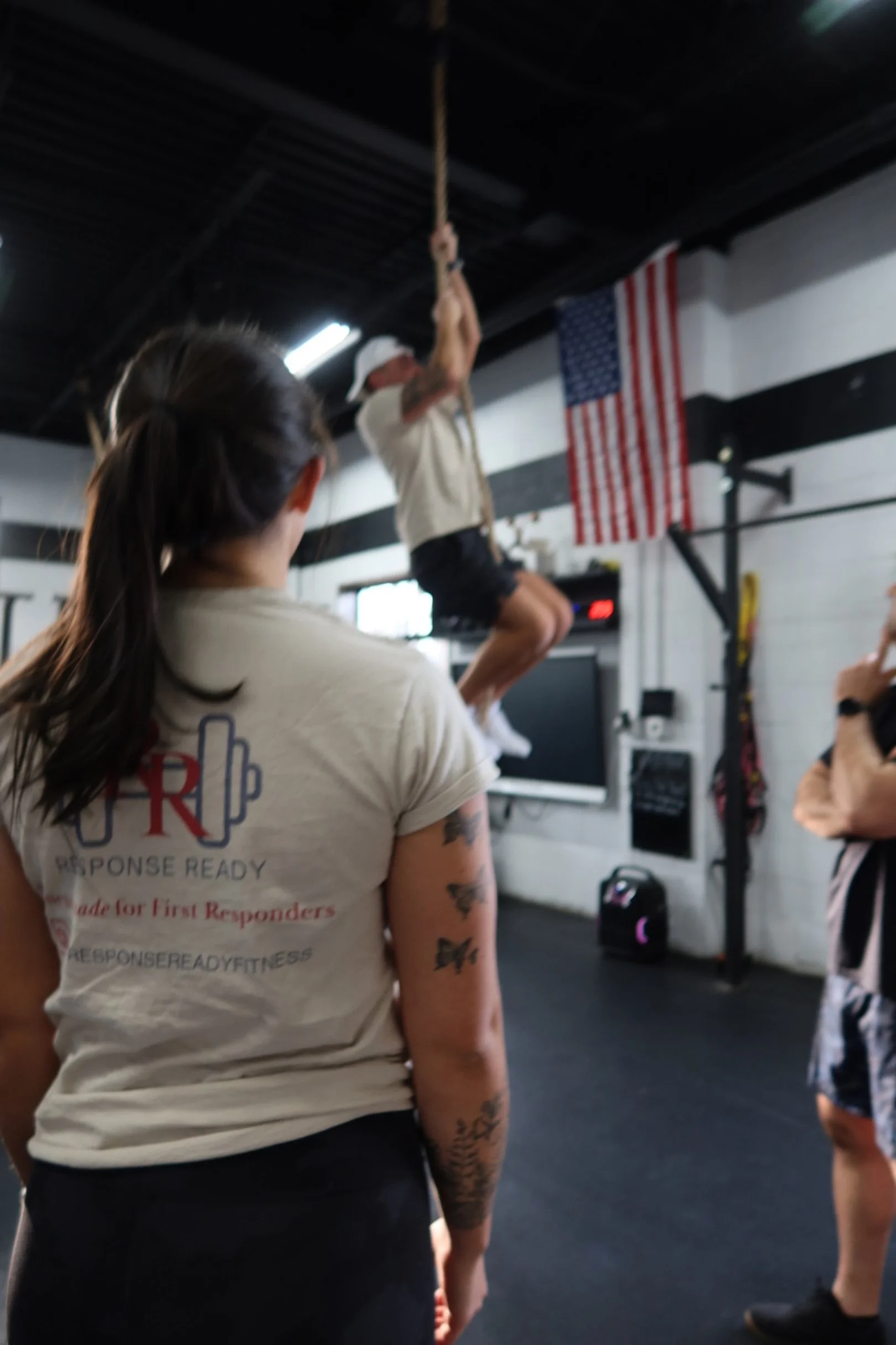 Person climbing a rope in a gym with a crowd watching, American flag on the wall.