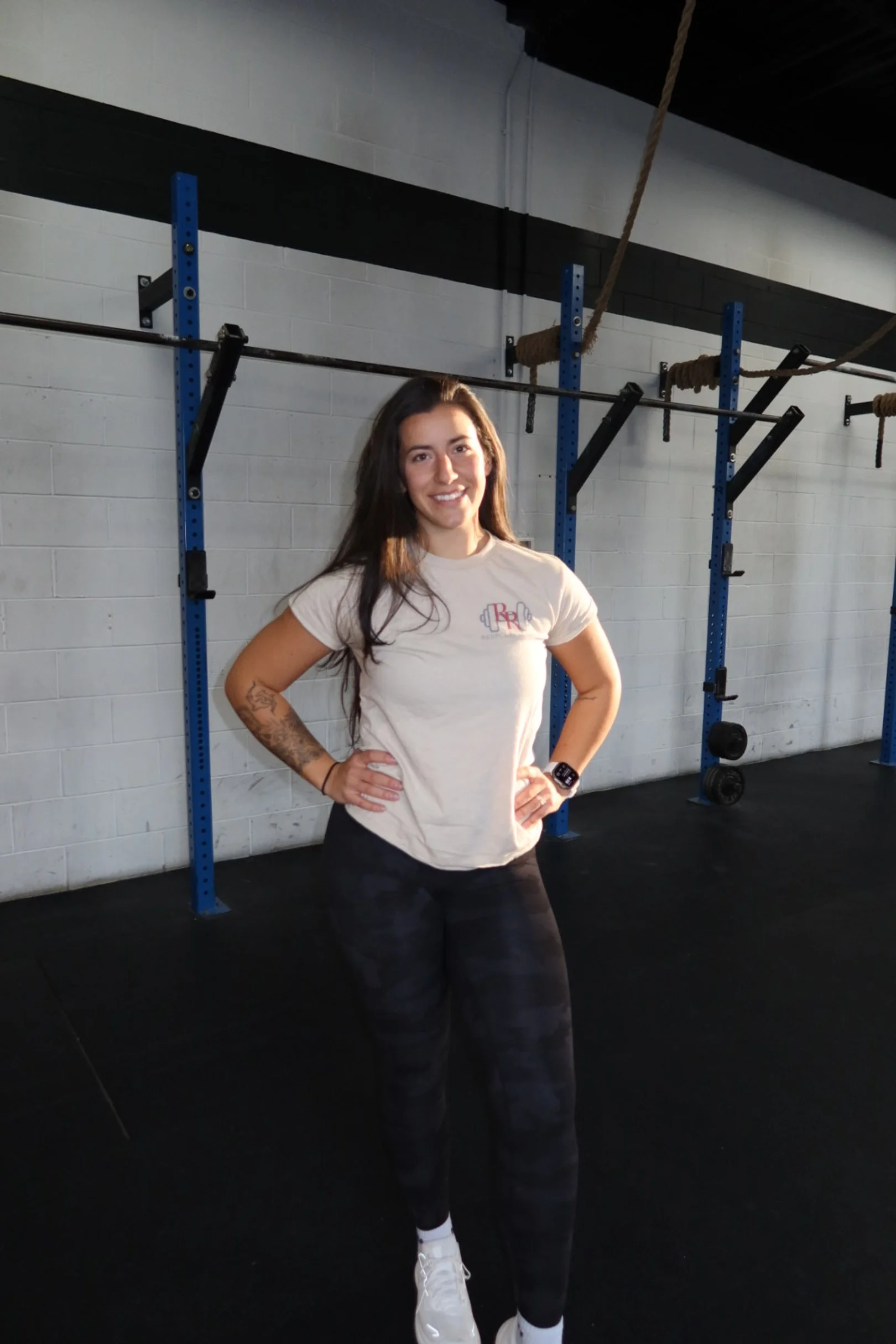 A woman standing in a gym with hands on her hips, smiling, wearing a beige t-shirt, black patterned leggings, and white sneakers.