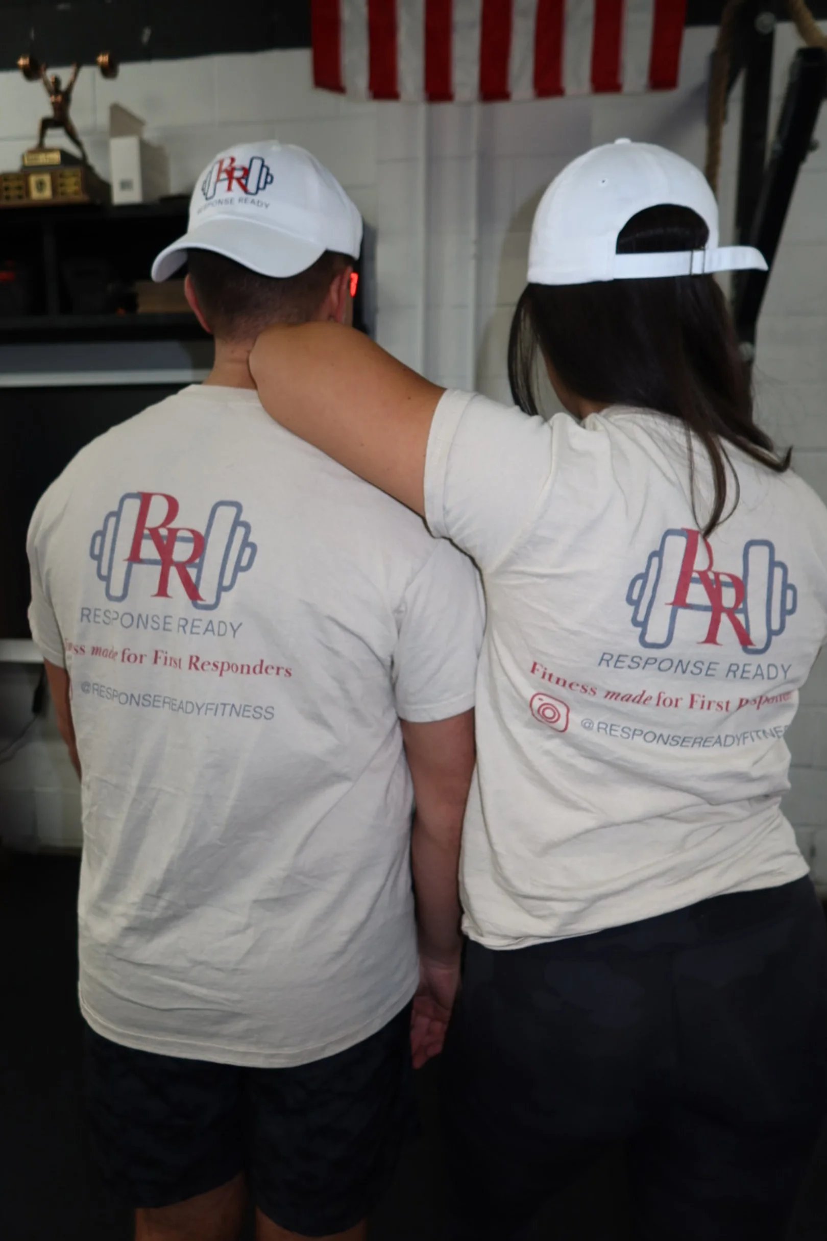 Two people with their backs to the camera wearing matching white t-shirts and white baseball caps, in a gym setting. Their shirts display a logo with the initials 'RR' and text reading 'Response Ready' and 'Fitness made for First Responders'.