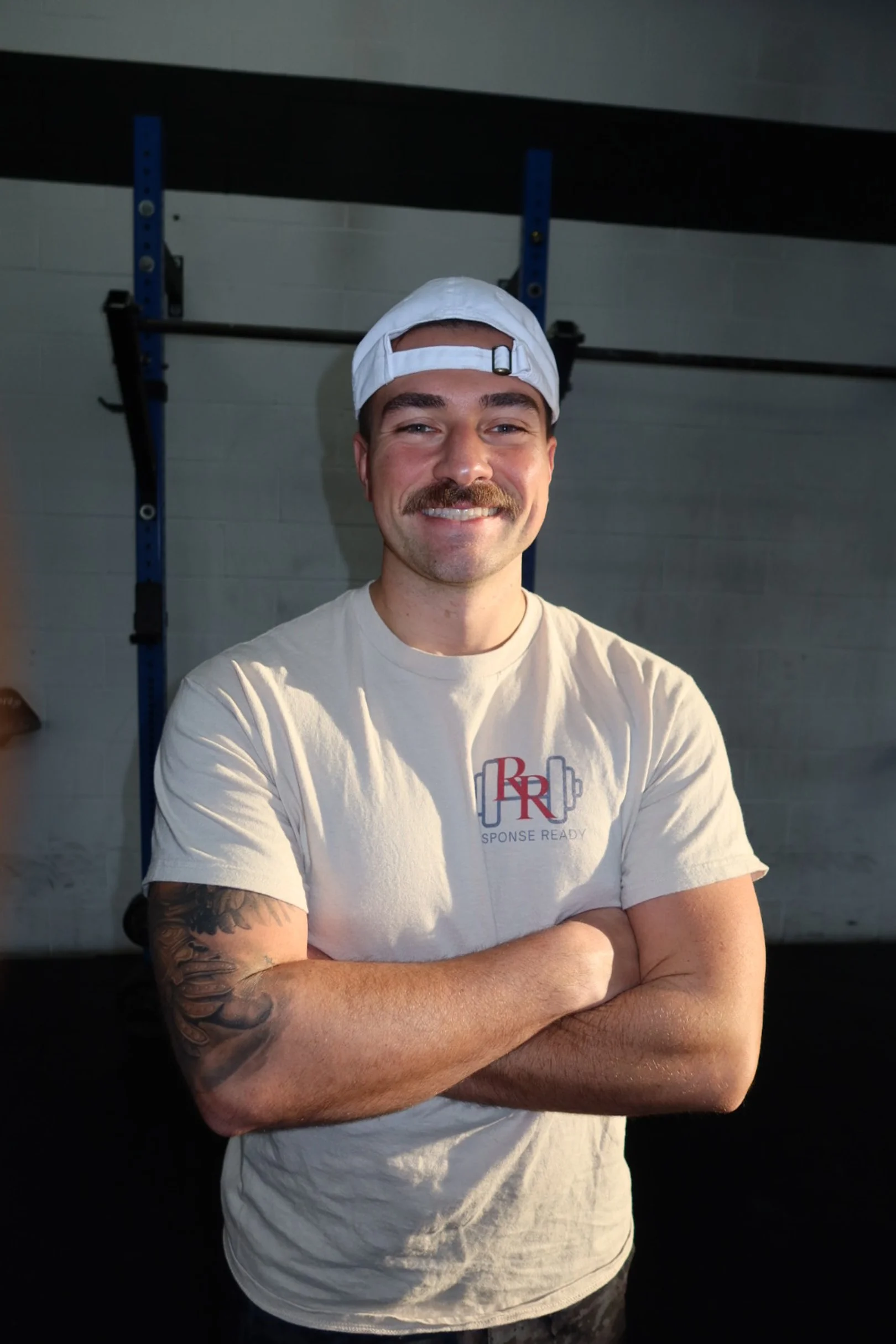 A man with a tattooed arm, wearing a white baseball cap backwards and a beige T-shirt with a logo, smiling with arms crossed in a gym.
