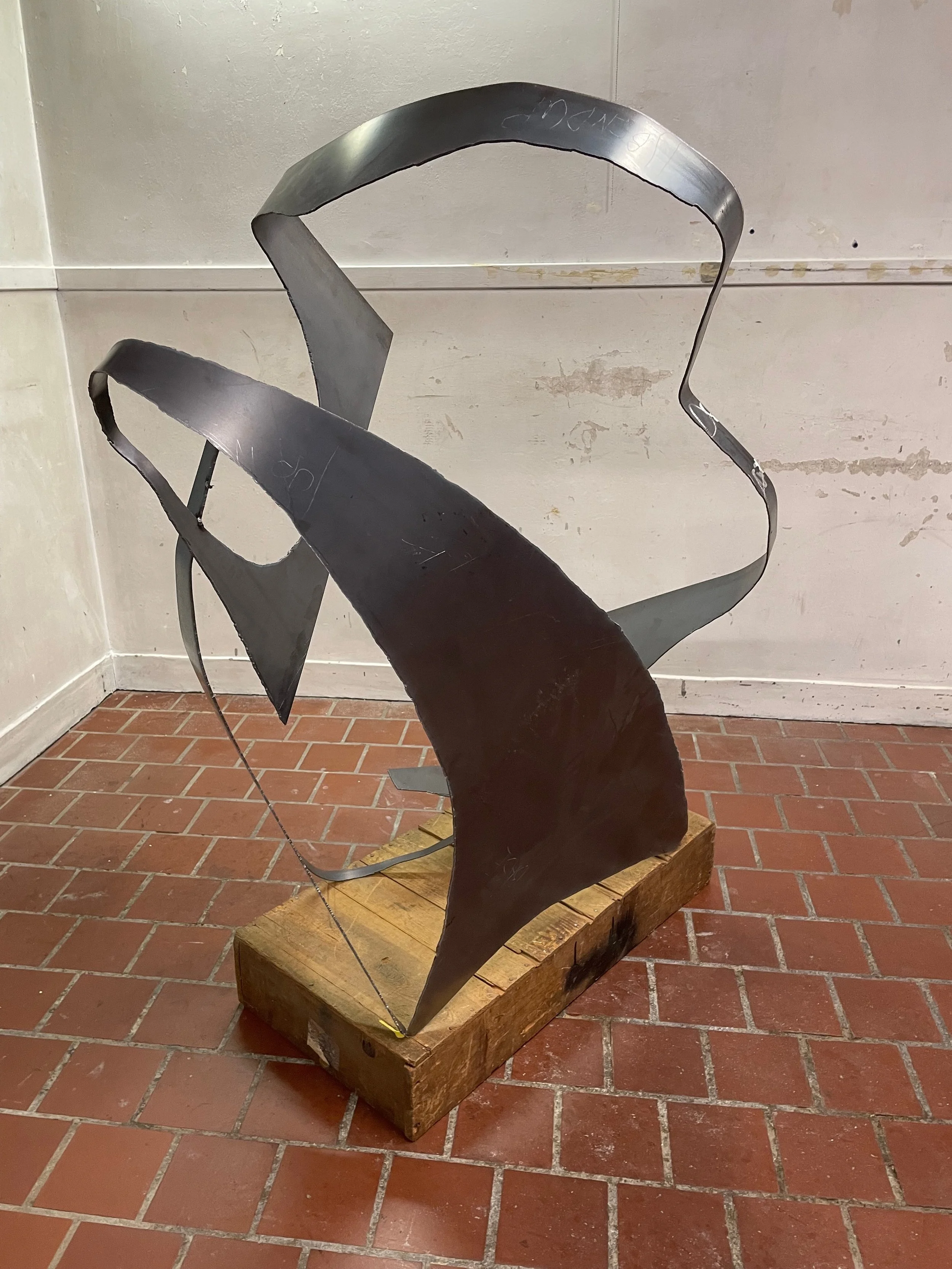 A modern abstract sculpture made of black metal, featuring curved and angular shapes, mounted on a wooden base, displayed indoors on a brick floor.