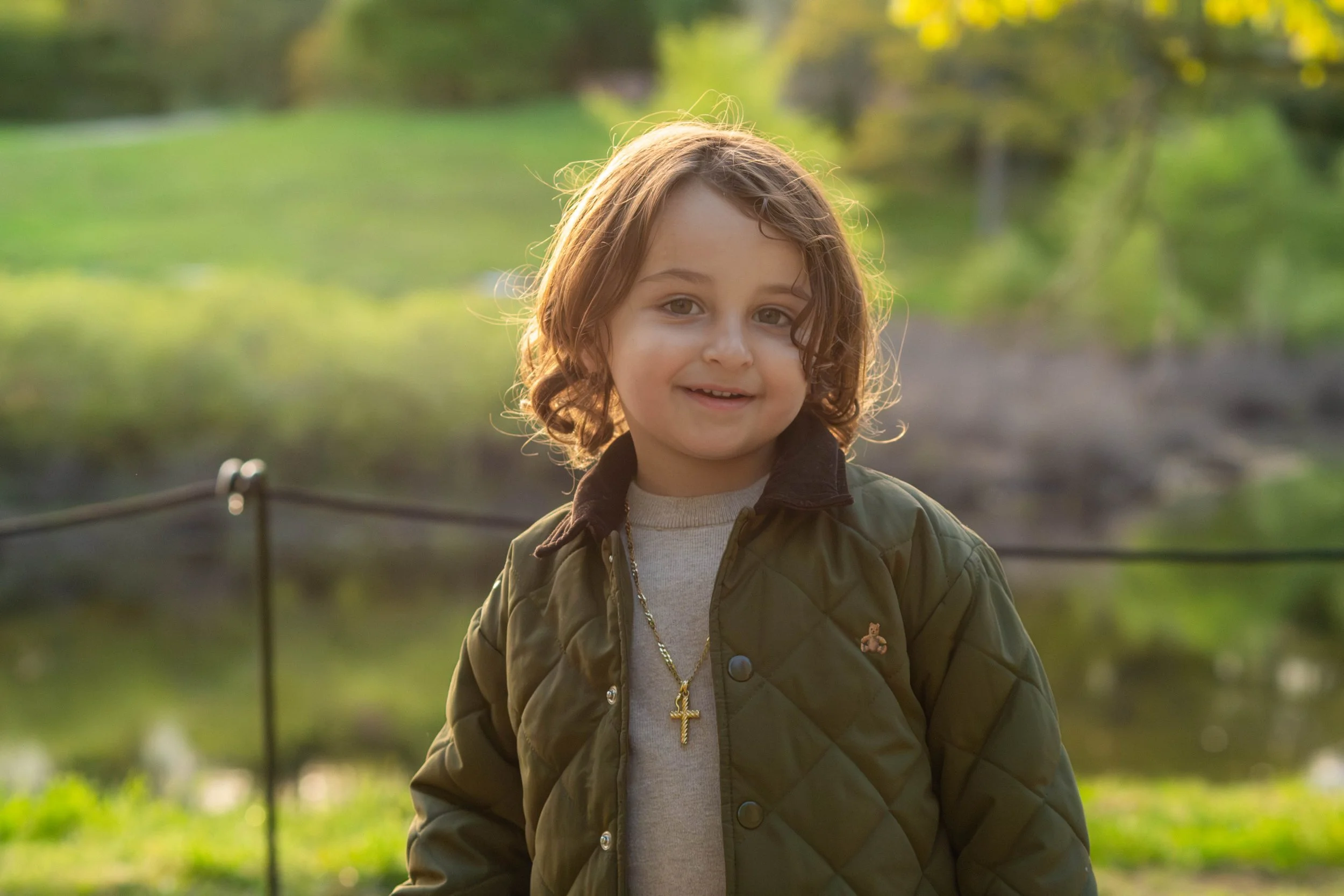A young girl with curly brown hair, wearing a green quilted jacket, a cross necklace, and a light gray shirt, standing outdoors in a park with blurred green trees and grass in the background, smiling at the camera.