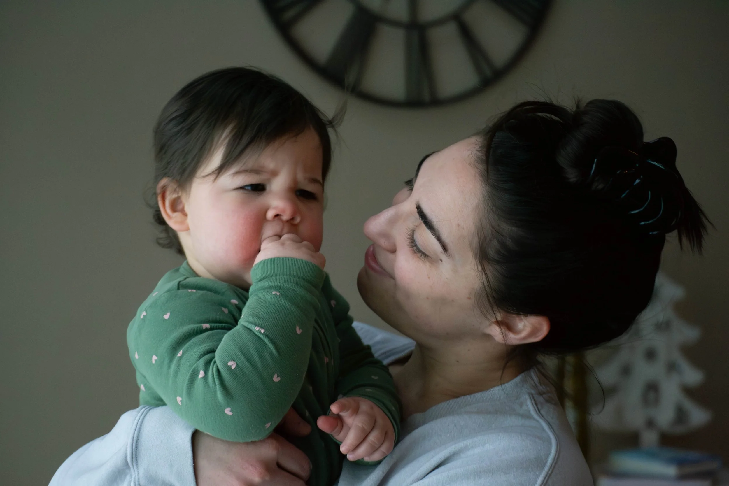 A woman holding a young child close, both smiling, with a clock on the wall in the background.
