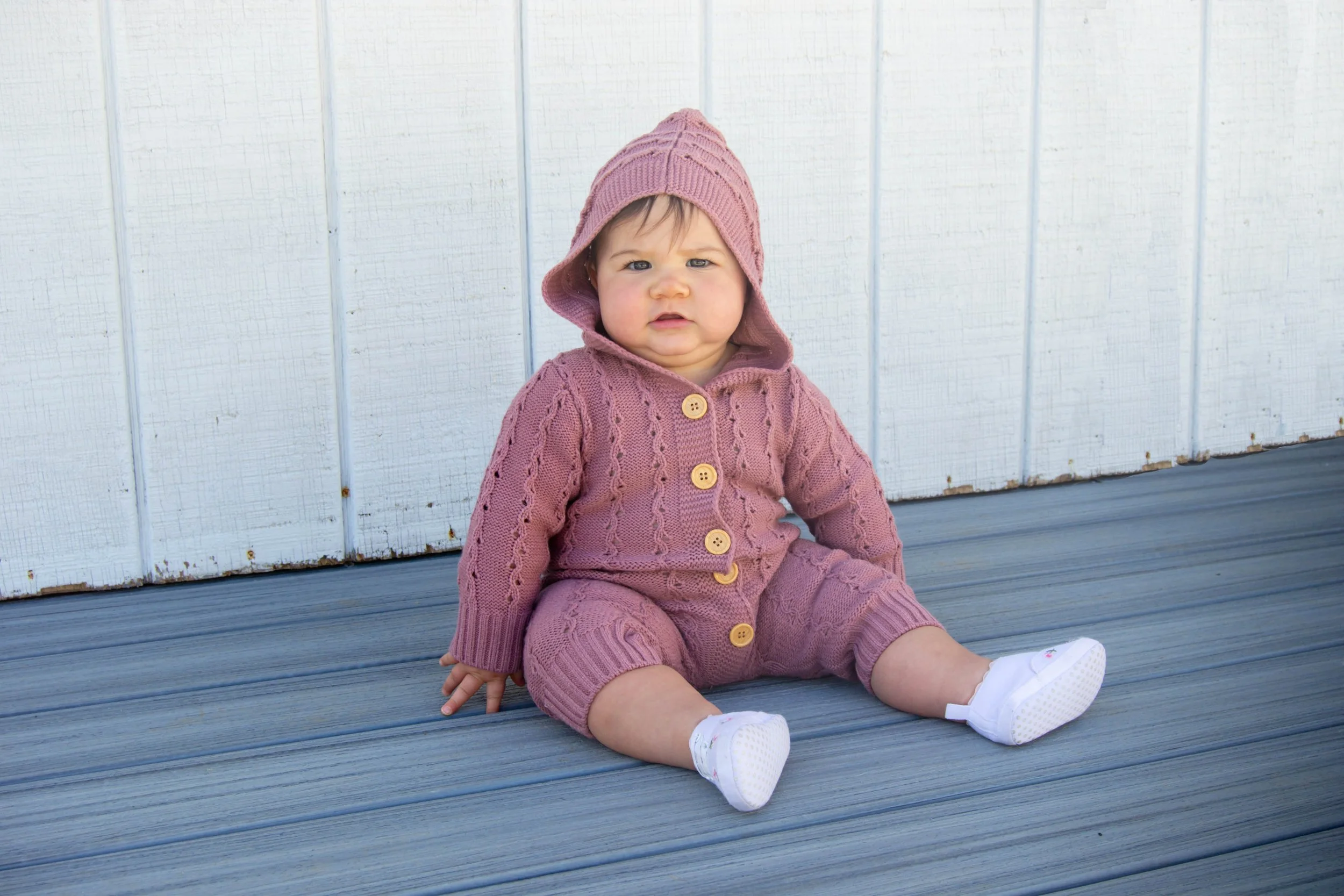 A baby girl sitting on a wooden deck, wearing a pink knit hoodie and matching pink pants, with white shoes and socks, in front of a white wooden wall.