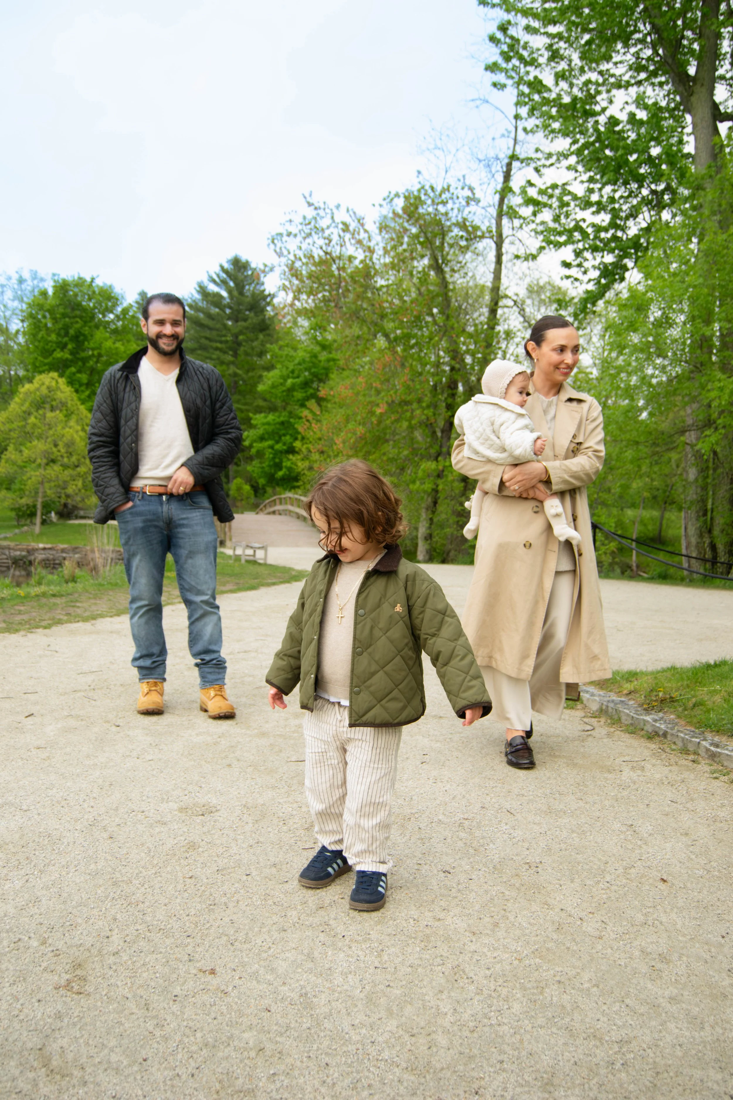 A family of four walking on a gravel path in a park with green trees. The father is smiling and wearing a black quilted jacket and jeans. The mother is holding a small child, and she is wearing a beige trench coat. The small child she is holding is d