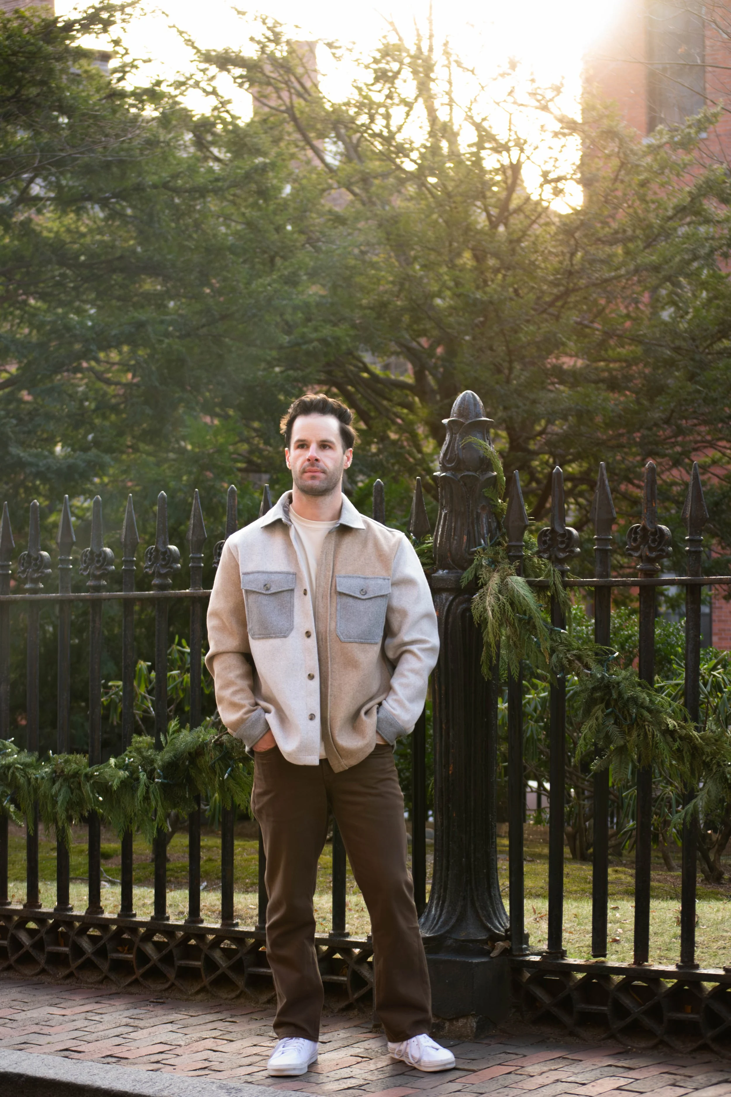 A man standing outdoors in front of a black iron fence with greenery and trees behind him, bathed in late afternoon sunlight.