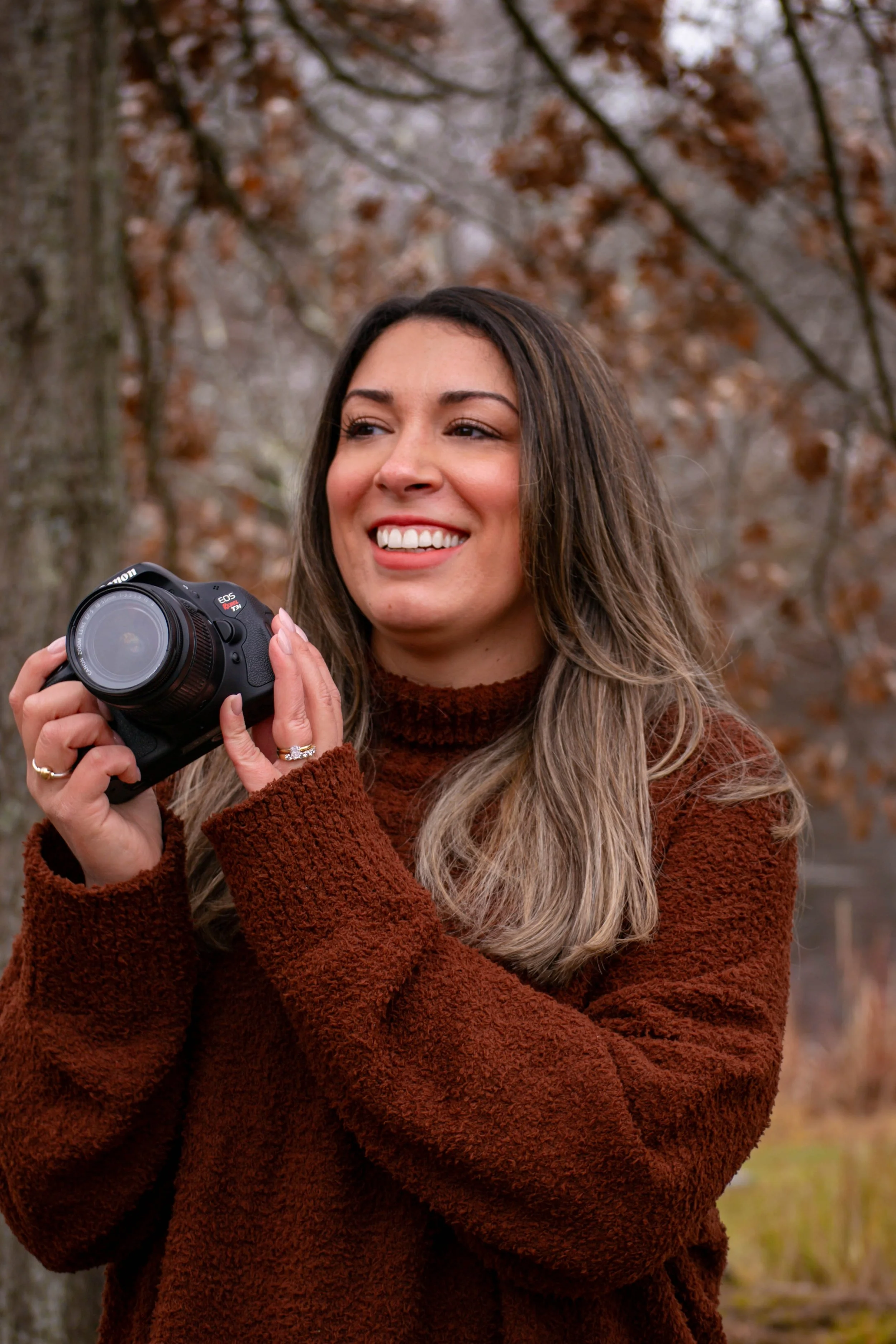 A woman holding a Canon camera outdoors during fall, smiling and looking to the side with brown leaves on trees in the background.