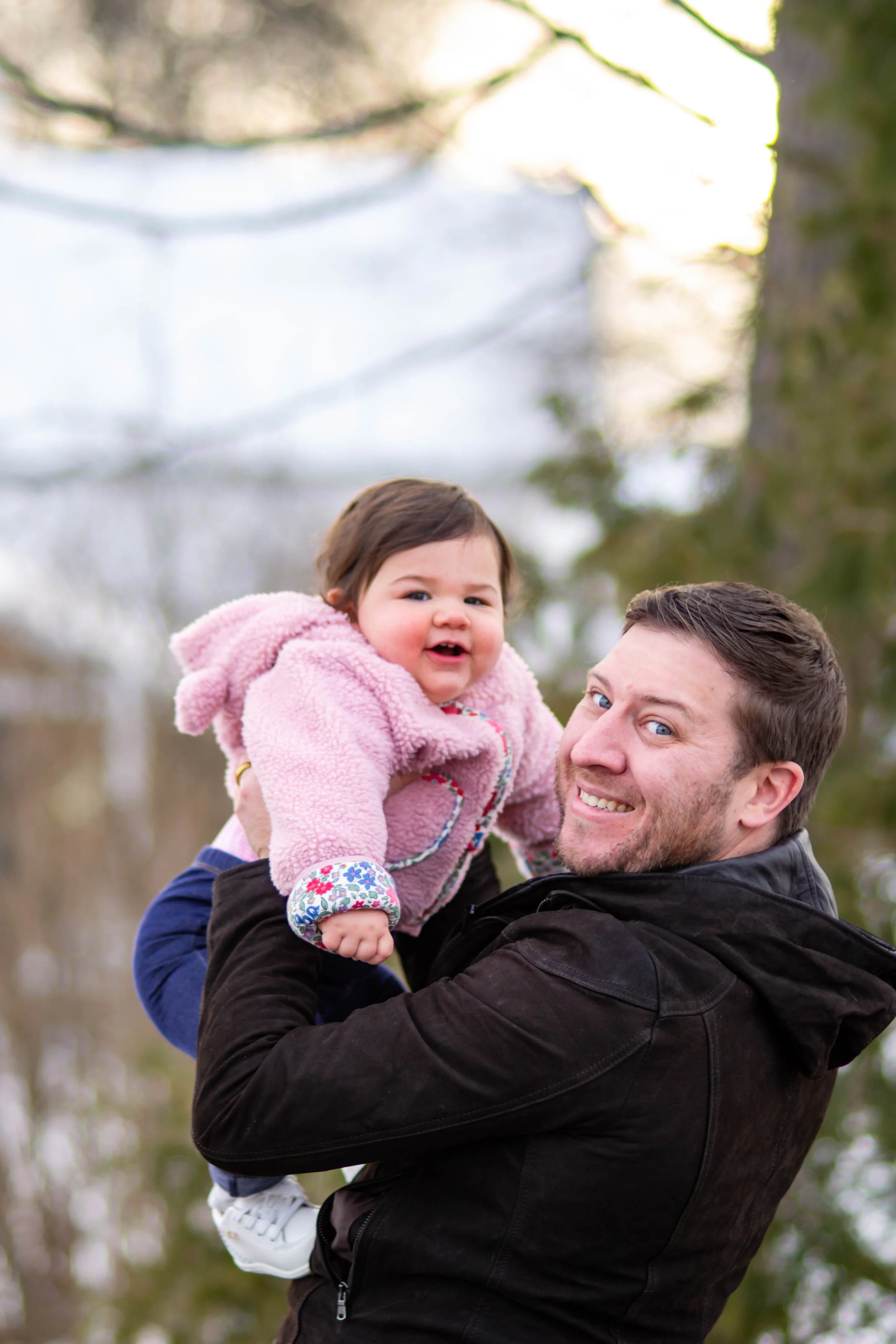 A man holding a smiling toddler girl wearing a pink fuzzy coat outdoors in a park with trees.