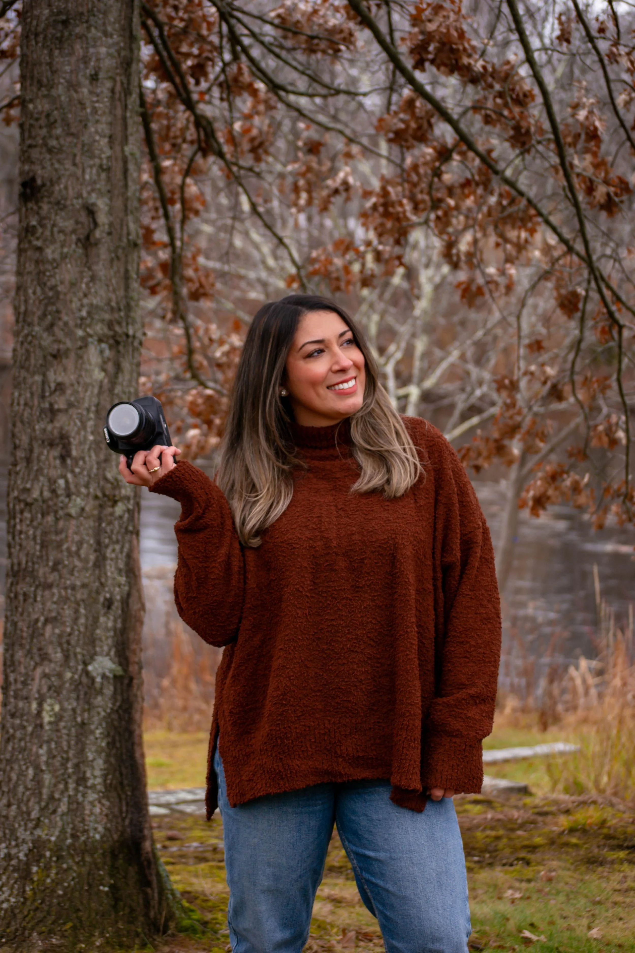 A woman standing outdoors near a tree with autumn leaves, smiling and holding a camera in her right hand.