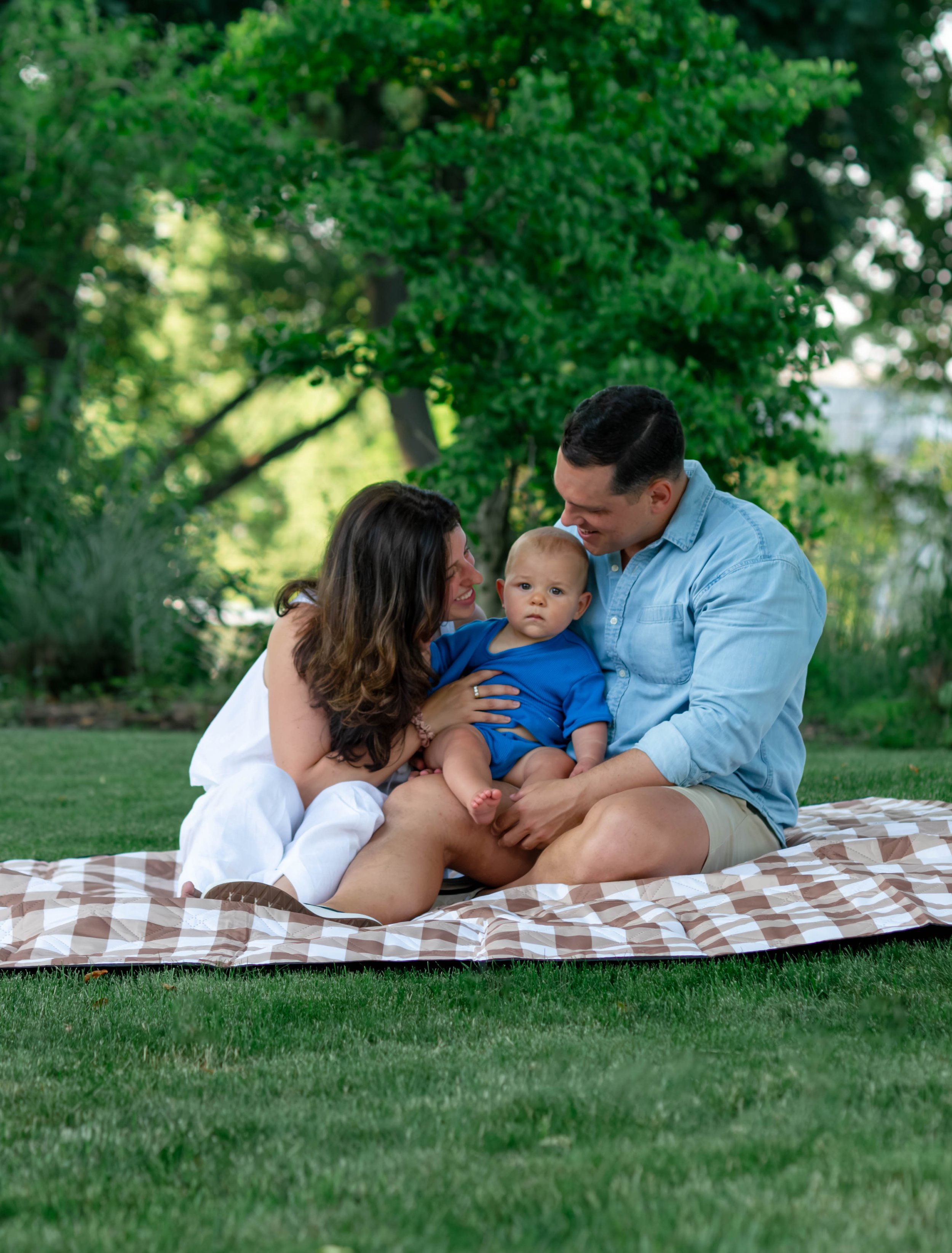 Family of three sitting on a blanket in a park, surrounded by green trees, with a woman, man, and a baby.