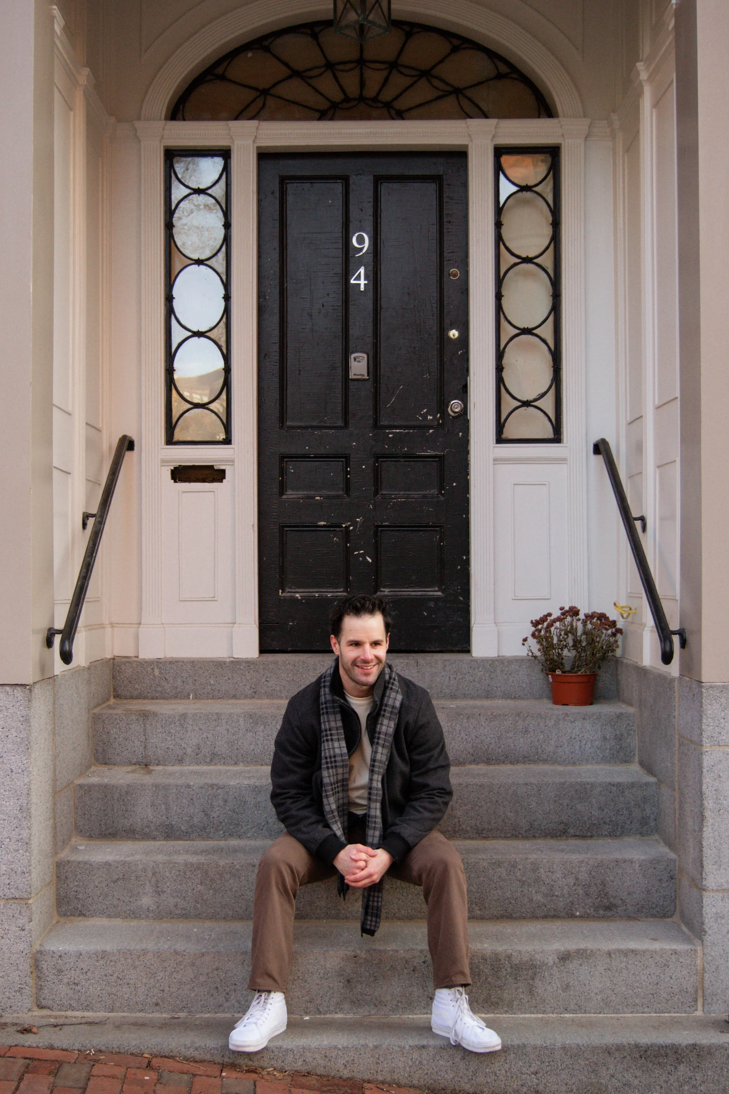 A man with dark hair and light skin, wearing a black jacket, beige pants, white sneakers, and a plaid scarf, sitting on concrete steps in front of a black front door with the number 94, flanked by two decorative windows, with a potted plant to the ri
