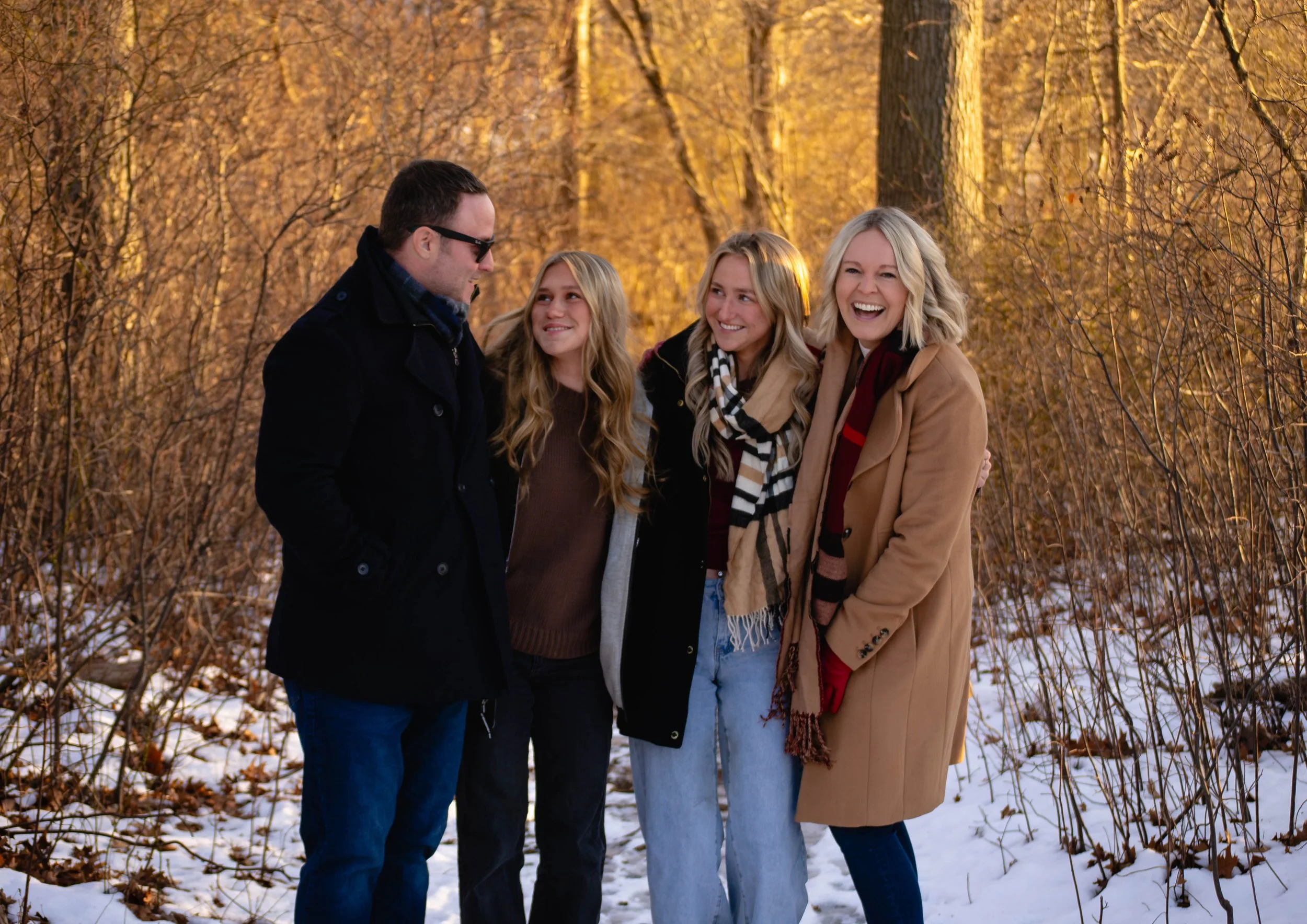 A group of four people, two women and two men, standing outdoors in a wooded area with fall foliage, smiling and enjoying each other's company.