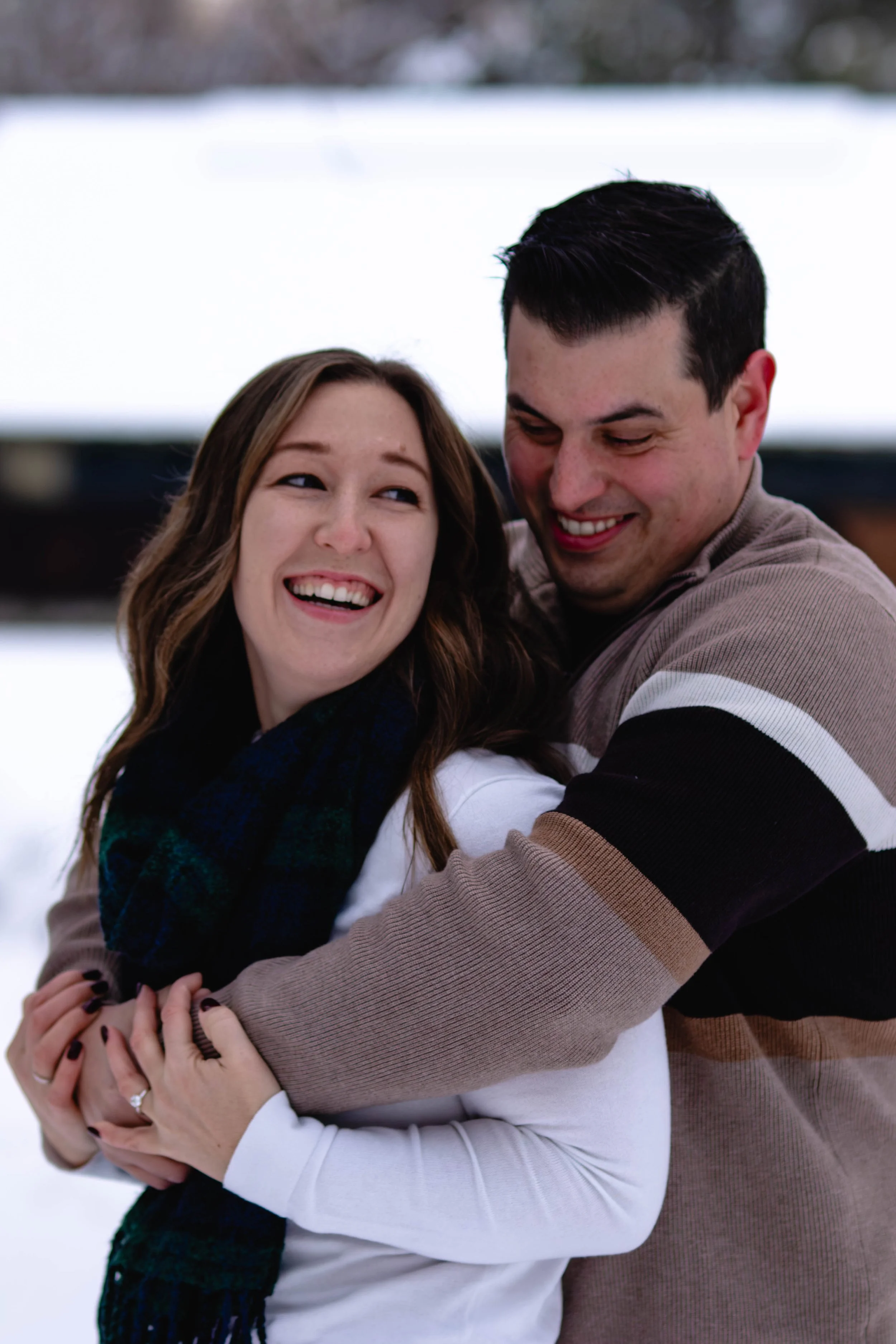 A couple is embracing outdoors with snow in the background. They are smiling and looking at each other, sharing a joyful moment.