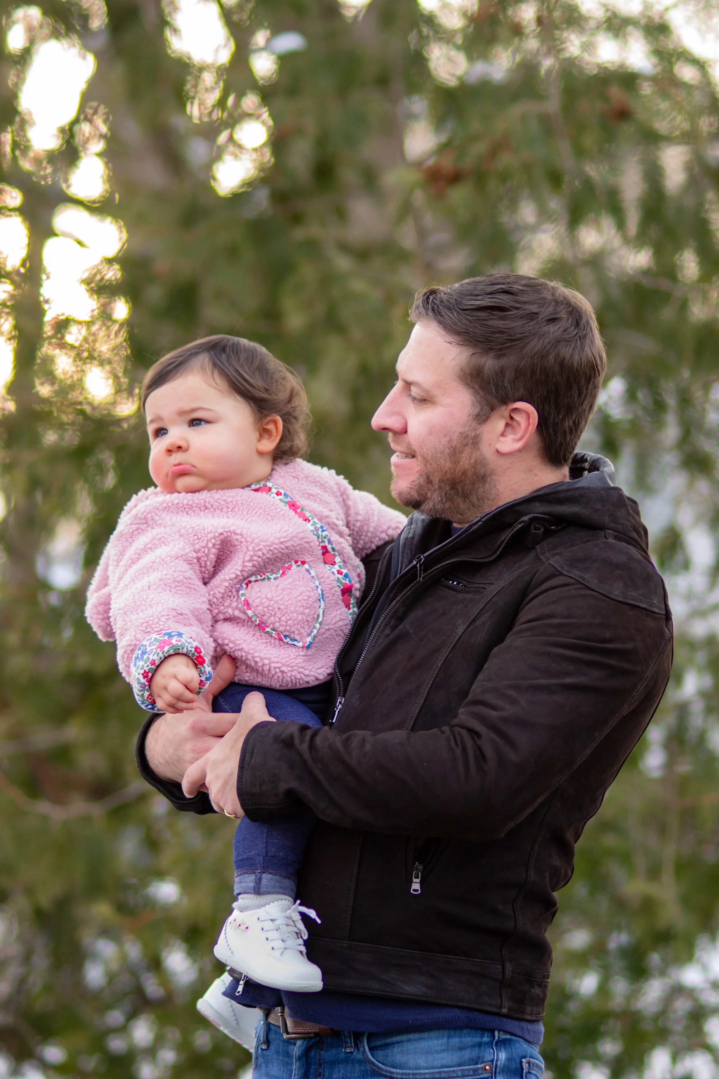A man holding a young girl outdoors, with trees in the background. The girl is wearing a pink fuzzy jacket with a heart design and looks thoughtful, while the man is smiling and looking at her.