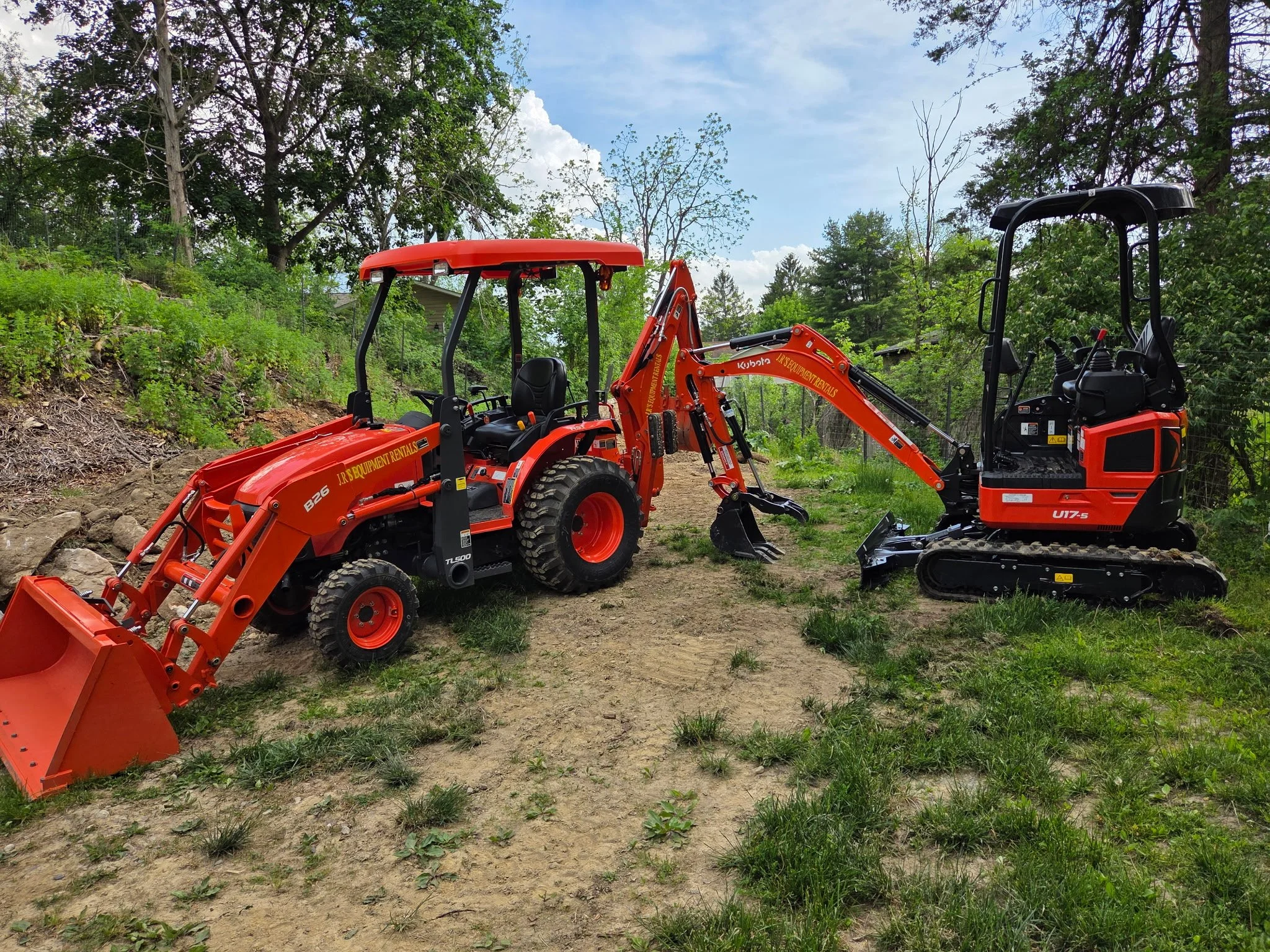 A bright orange backhoe loader and a mini excavator parked on dirt ground outdoors surrounded by green trees and bushes with a partly cloudy sky overhead.