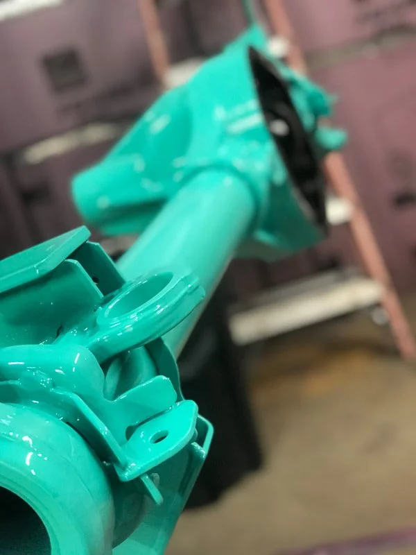 Close-up of a turquoise or teal painted metal pipe or tube, with industrial equipment or part in the background.