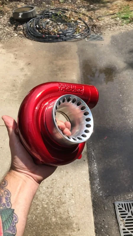 Person holding a red turbocharger in their left hand, with a garage or workshop area in the background, including black coiled hoses and an air filter on the ground.