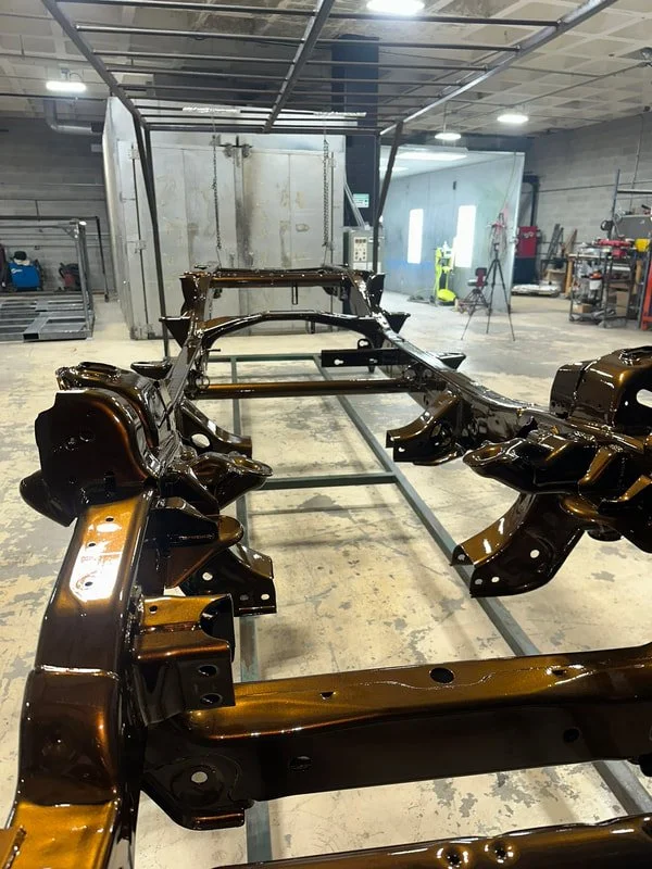 An automobile chassis painted in a shiny, metallic brown, inside an industrial workshop with tools and equipment in the background.