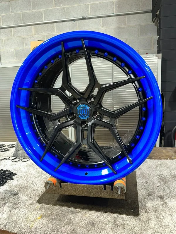 A custom car wheel with a black five-spoke rim and a blue illuminated outer ring, mounted on a foam and wood stand in a workshop setting.