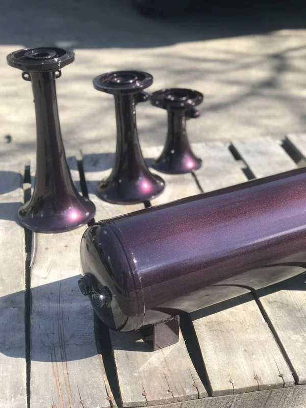 Three iridescent metal objects resembling small vases or sculptures and a cylindrical object lying on a wooden bench outside on a sunny day.