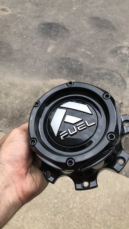 A person's hand holding a black fuel cap with the logo and the word 'FUEL' on it, against a concrete ground background.
