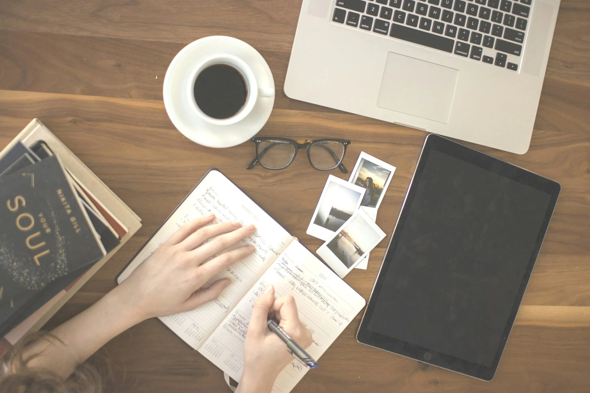 A top-down view of a workspace with a laptop, a tablet, a notebook, a pair of glasses, a cup of black coffee, an open book, and some Polaroid pictures on a wooden desk.