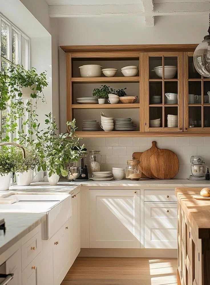 Sunlit kitchen with open wooden shelves displaying white bowls, plates, and cups, white cabinetry, a wooden cutting board, potted plants by the window, and a white tile backsplash.