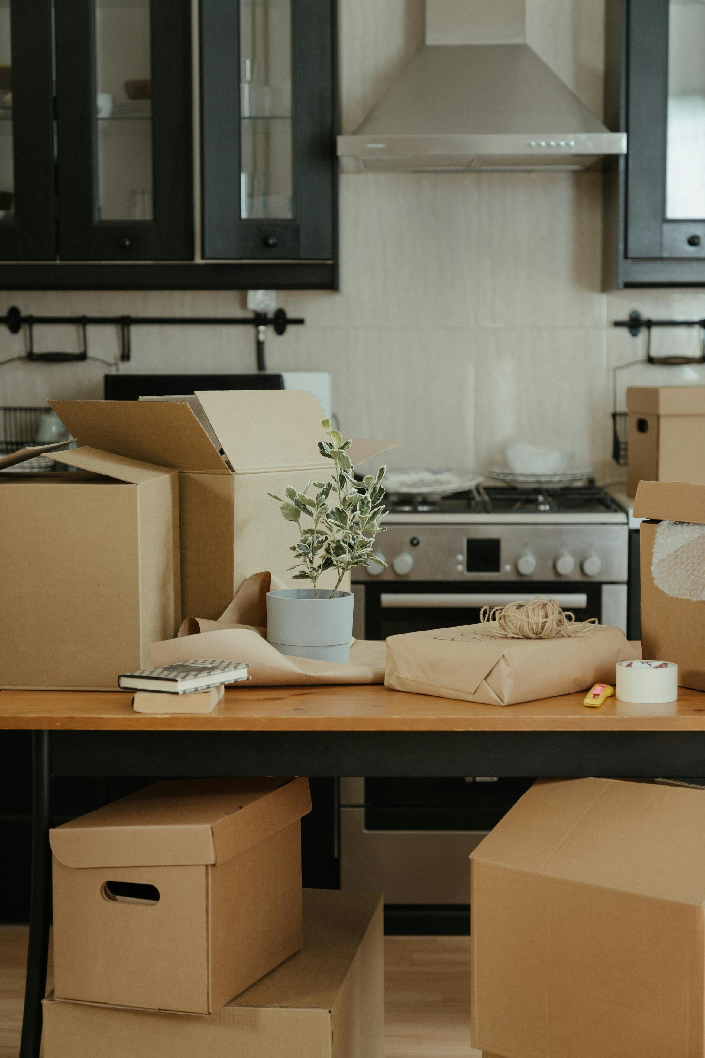 Kitchen with open cardboard boxes, a potted plant, wrapping paper, tape, and yarn on a table, with black cabinets, a stove, and a range hood in the background.