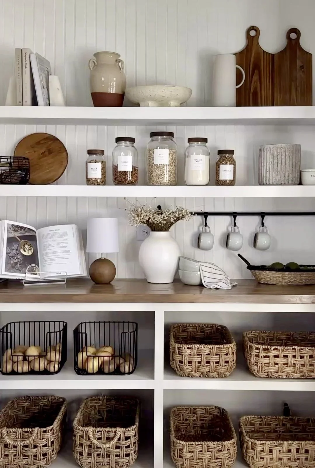 A tidy kitchen shelf with jars of grains and spices, a white vase with flowers, a small book open on a stand, and woven baskets on a lower shelf.