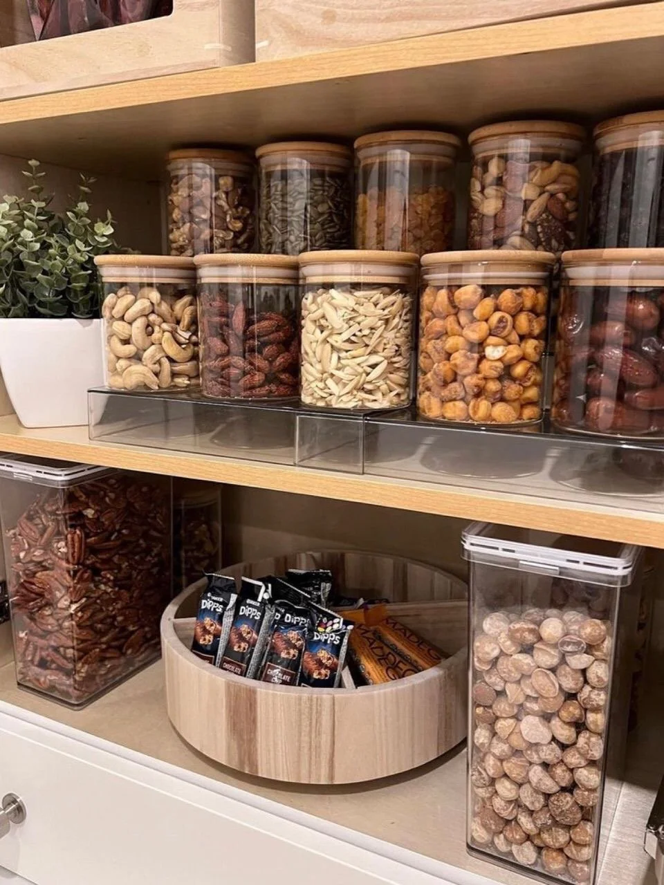 Various jars of mixed nuts and snack bars on a wood shelf, with a small potted plant on the left.