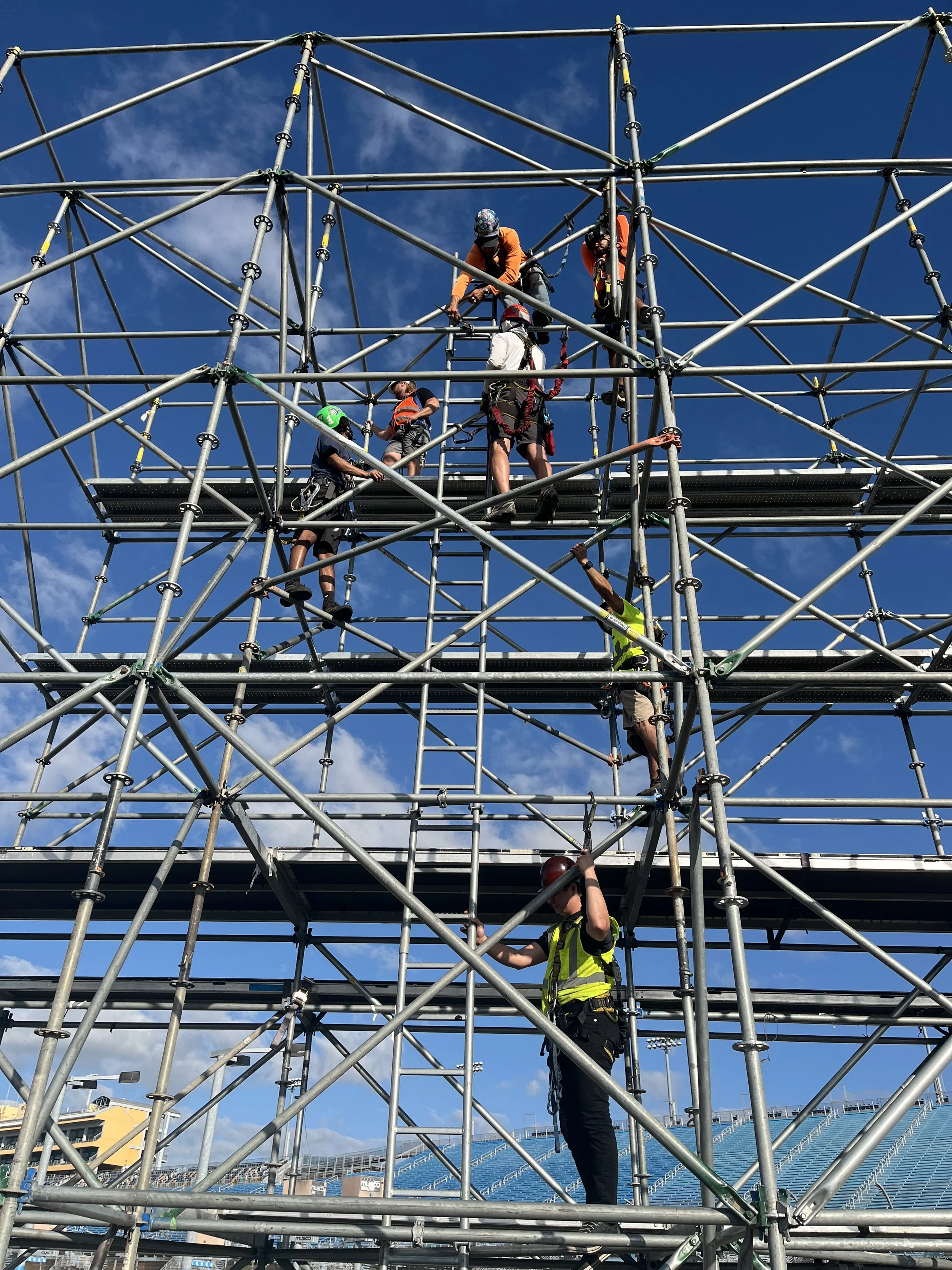 Workers in fall protection safety harness and gear work on large metal scaffold structure against a blue sky with clouds.