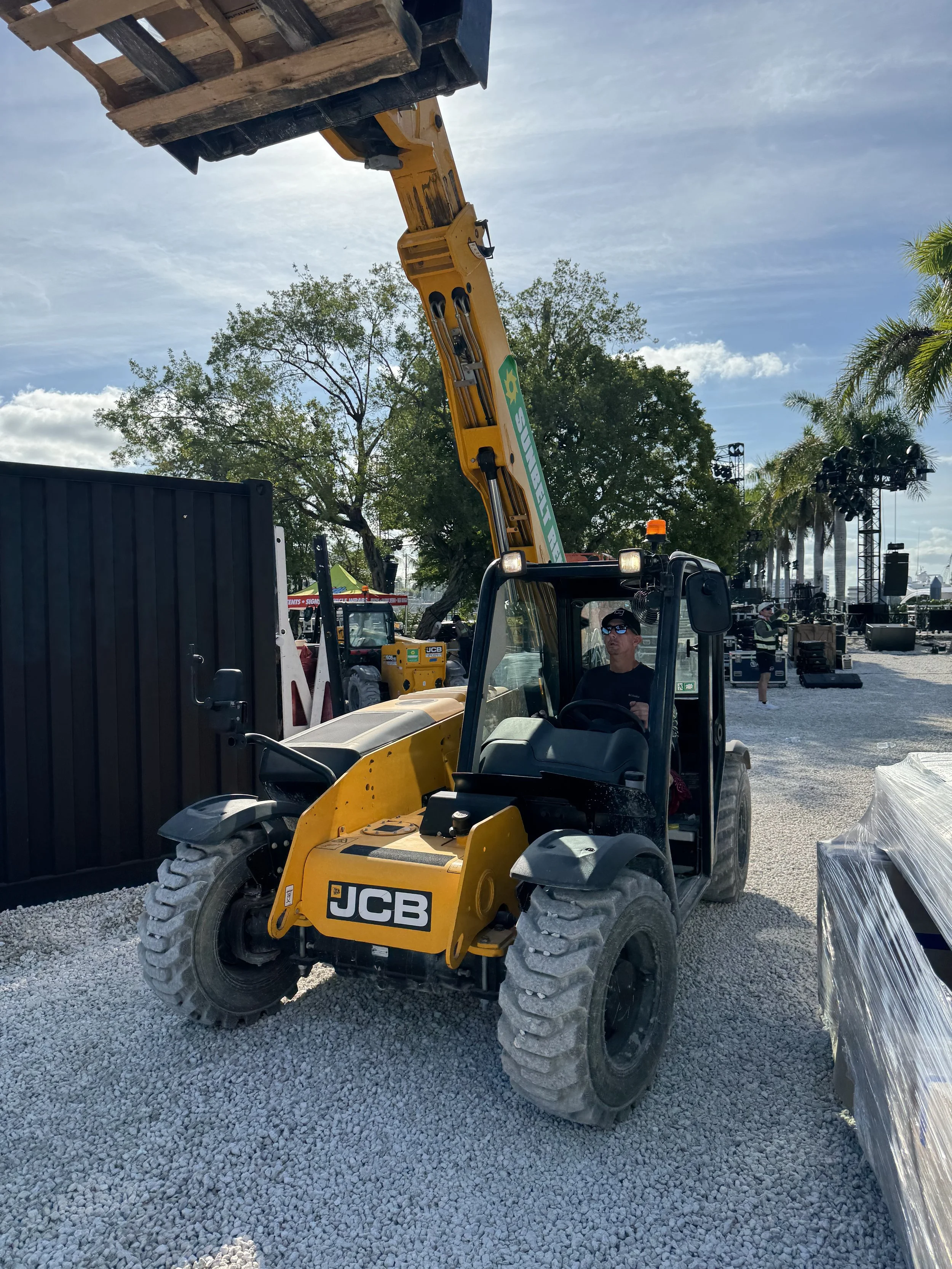 A person operating a JCB forklift lifting a wooden pallet at an outdoor event setup, with a stage, trees, and palm trees in the background.