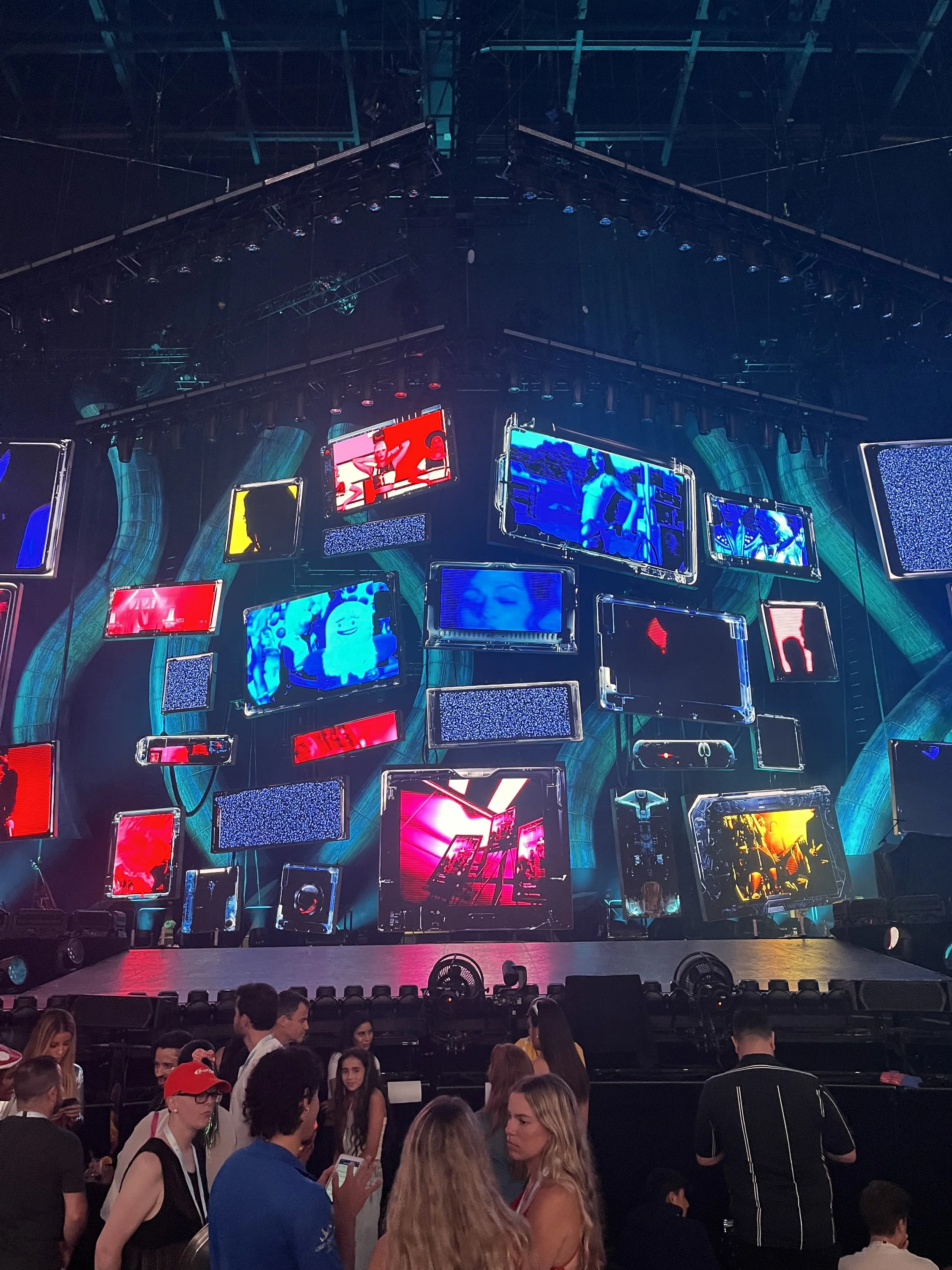 Concert stage with multiple screens displaying colorful visuals, and people gathered in front.
