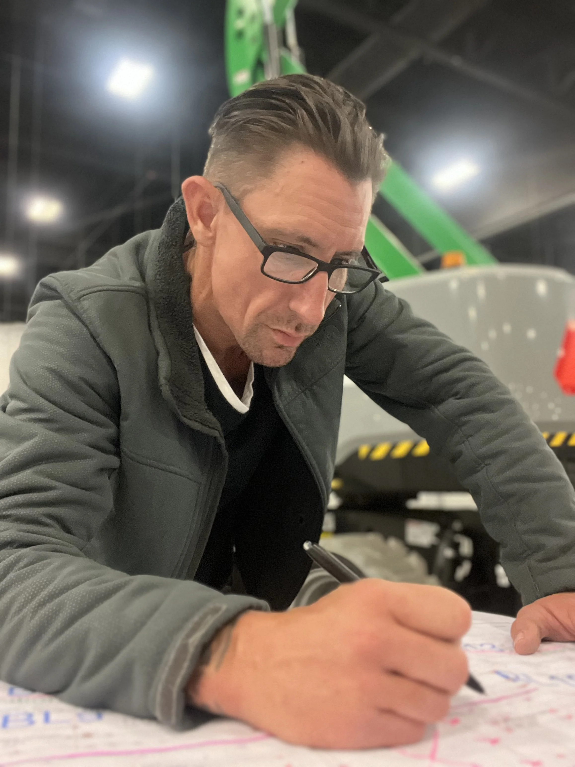 A man with glasses and a gray jacket signing or writing on a large sheet of paper.