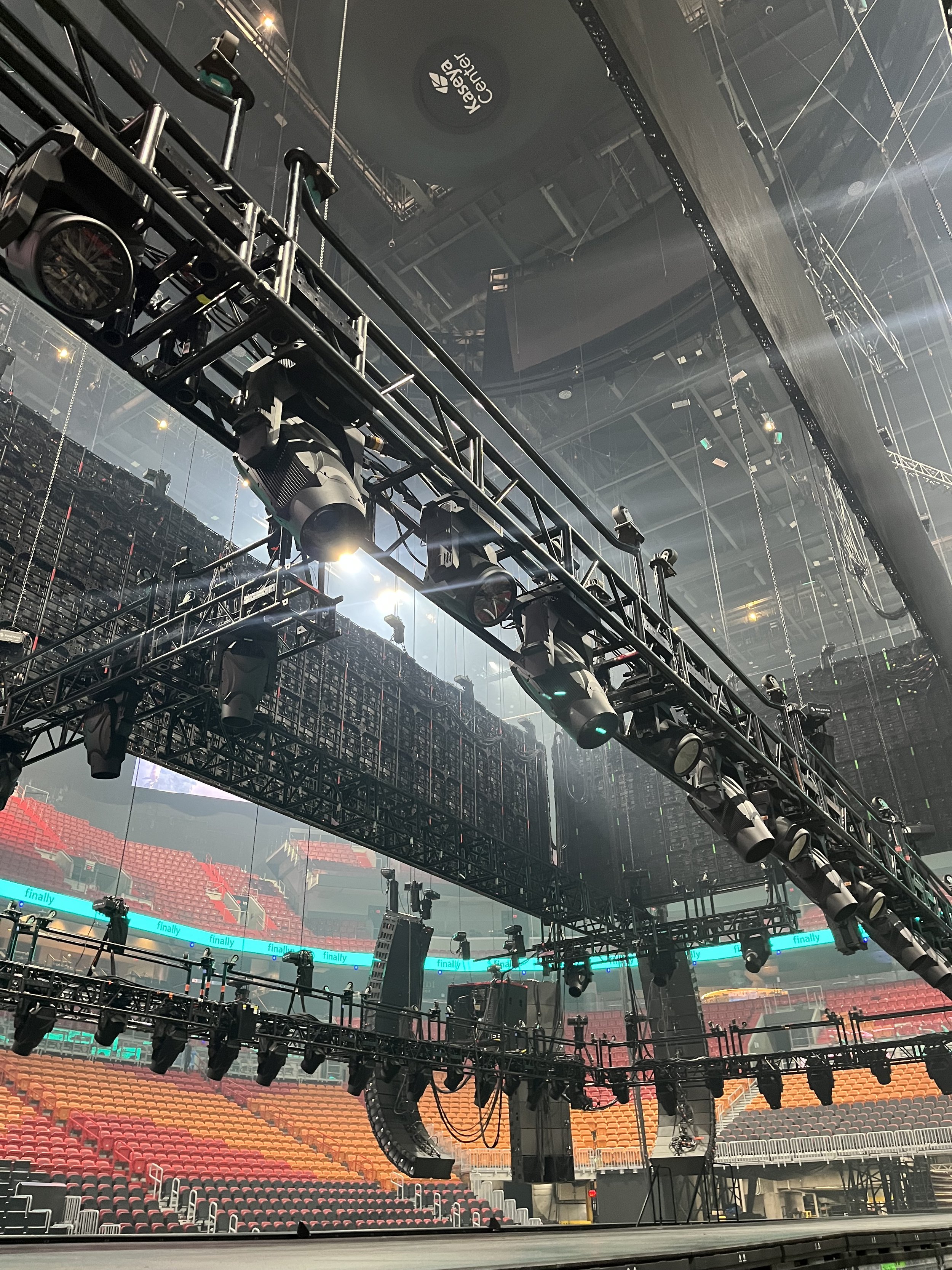 View of a large indoor arena or concert venue with stage lighting equipment and rigging hanging from the ceiling, empty seating in the background.